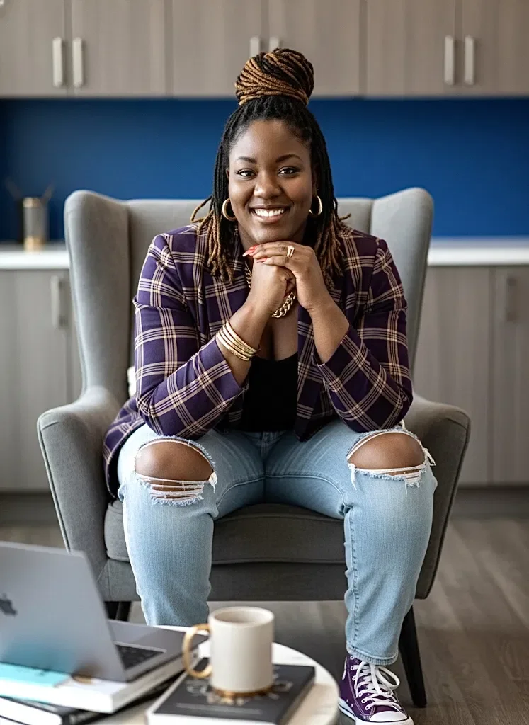 A woman sitting in a gray armchair, smiling, with her hands clasped. She has dreadlocks in a bun, wears a plaid blazer and ripped jeans. There is a laptop, a cup, and a notebook on a table in front of her.