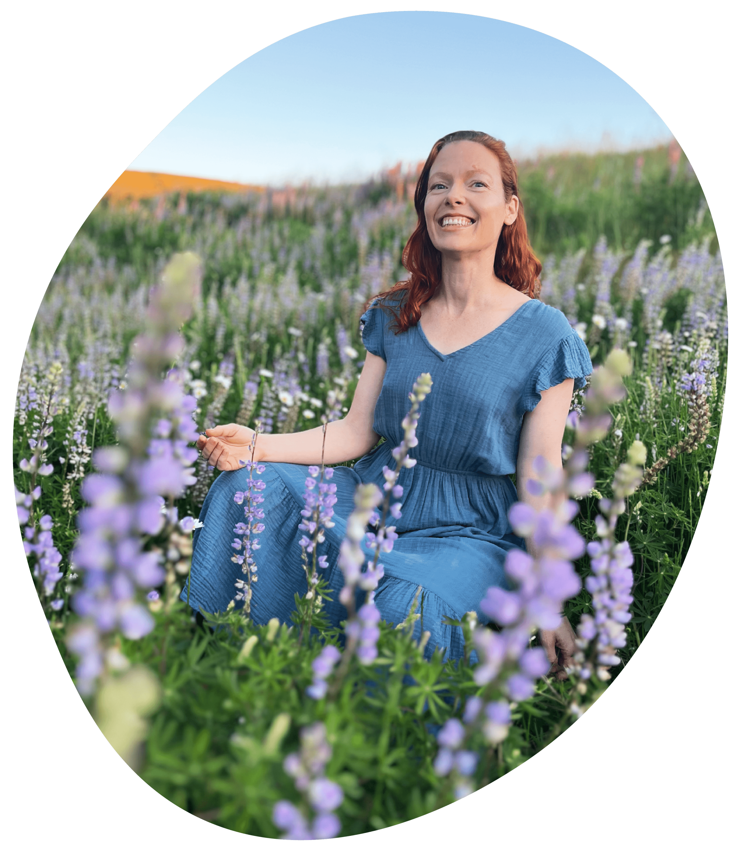 A woman with red hair sitting in a field of purple and white flowers, smiling, wearing a blue dress, with a blurred background of more flowers and a clear sky.