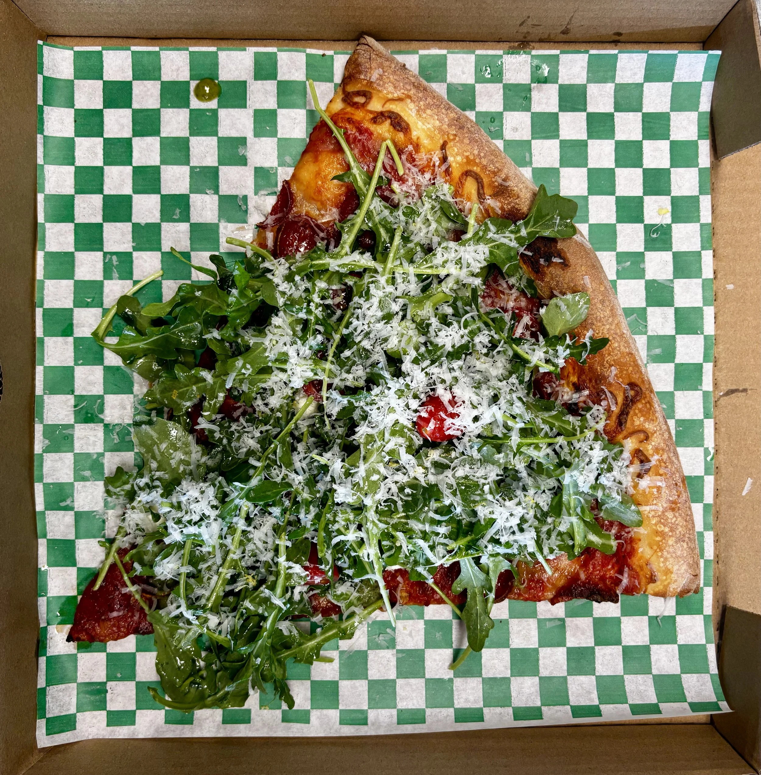 A slice of pepperoni pizza with leafy greens and shredded cheese inside a cardboard pizza box with green and white checkered paper.