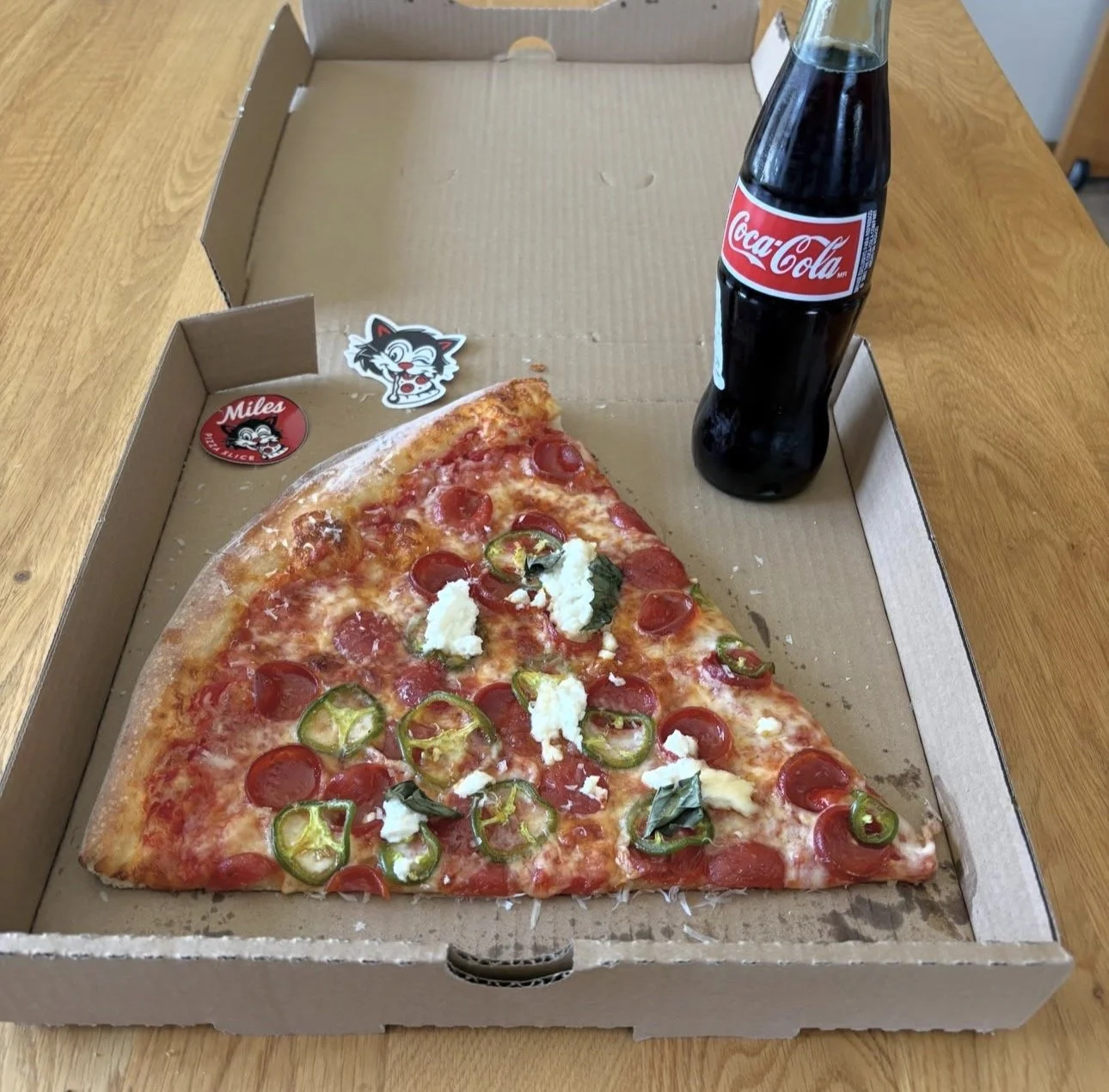 A slice of pepperoni and jalapeno pizza with ricotta, served in an open pizza box alongside a glass bottle of Coca-Cola on a wooden table.