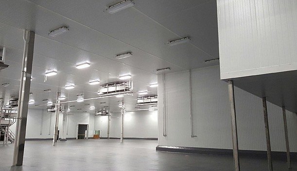 Empty, brightly lit industrial or warehouse space with white walls and ceiling, fluorescent lights, metal support poles, and a gray concrete floor.