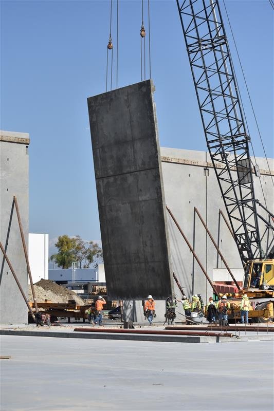 Construction site with a crane lifting a large concrete panel while workers guide it into position.