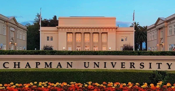 The entrance of Chapman University with a large sign displaying its name, surrounded by colorful flowers and historic buildings in the background.