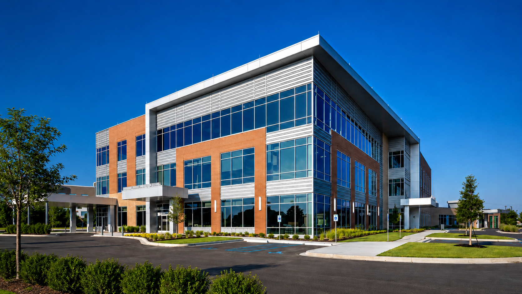 Modern office building with large glass windows, surrounded by parking lot and landscaping, on a sunny day.