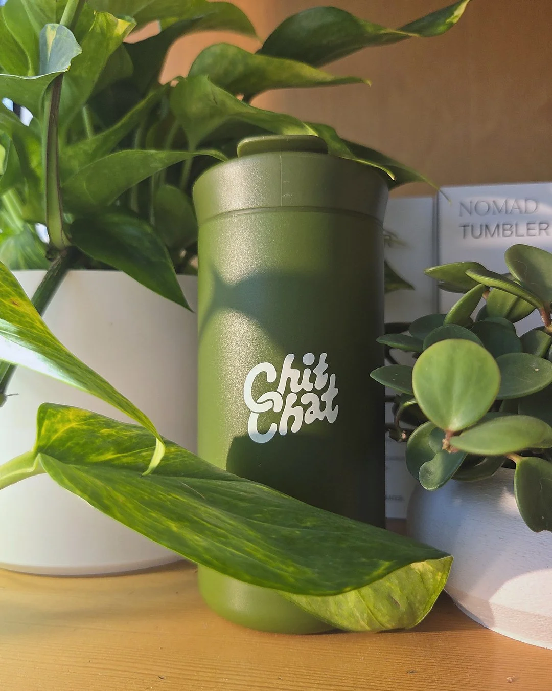 Green Chilly Chat tumbler among potted green plants on a wooden surface.