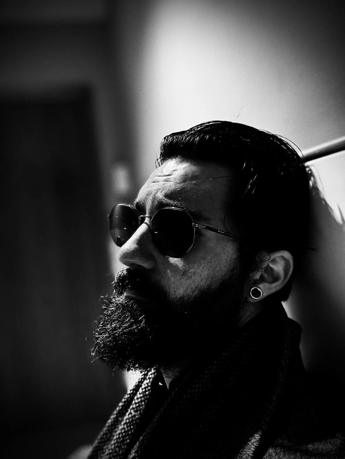 Black and white photo of a man with a beard and mustache wearing sunglasses and an earring, leaning against a wall with his eyes closed.