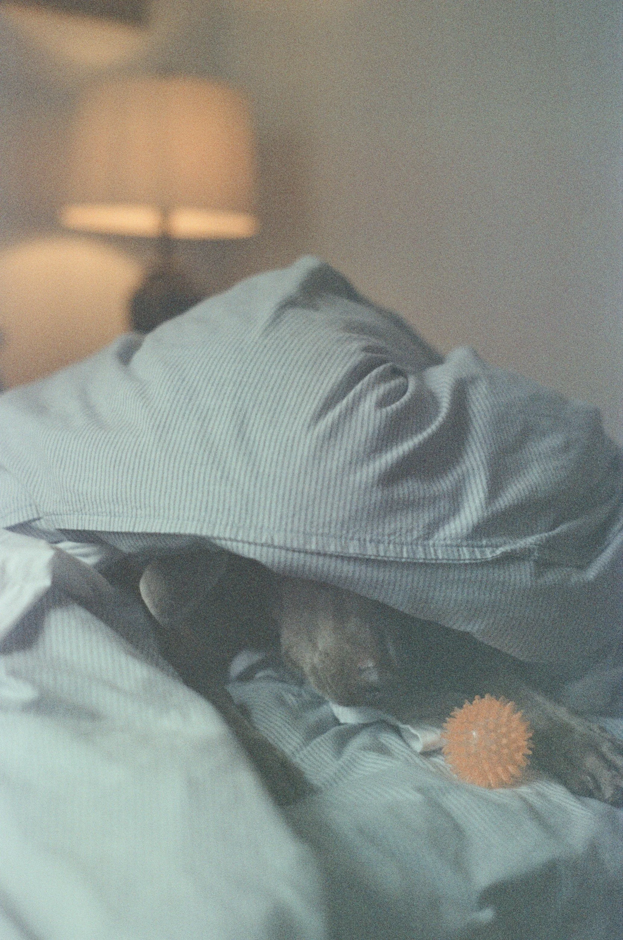 A close-up view of a person wearing gray and black striped pajamas, lying down with a grey blanket covering their body. There is a lamp in the background and a fuzzy orange object in the lower right corner.