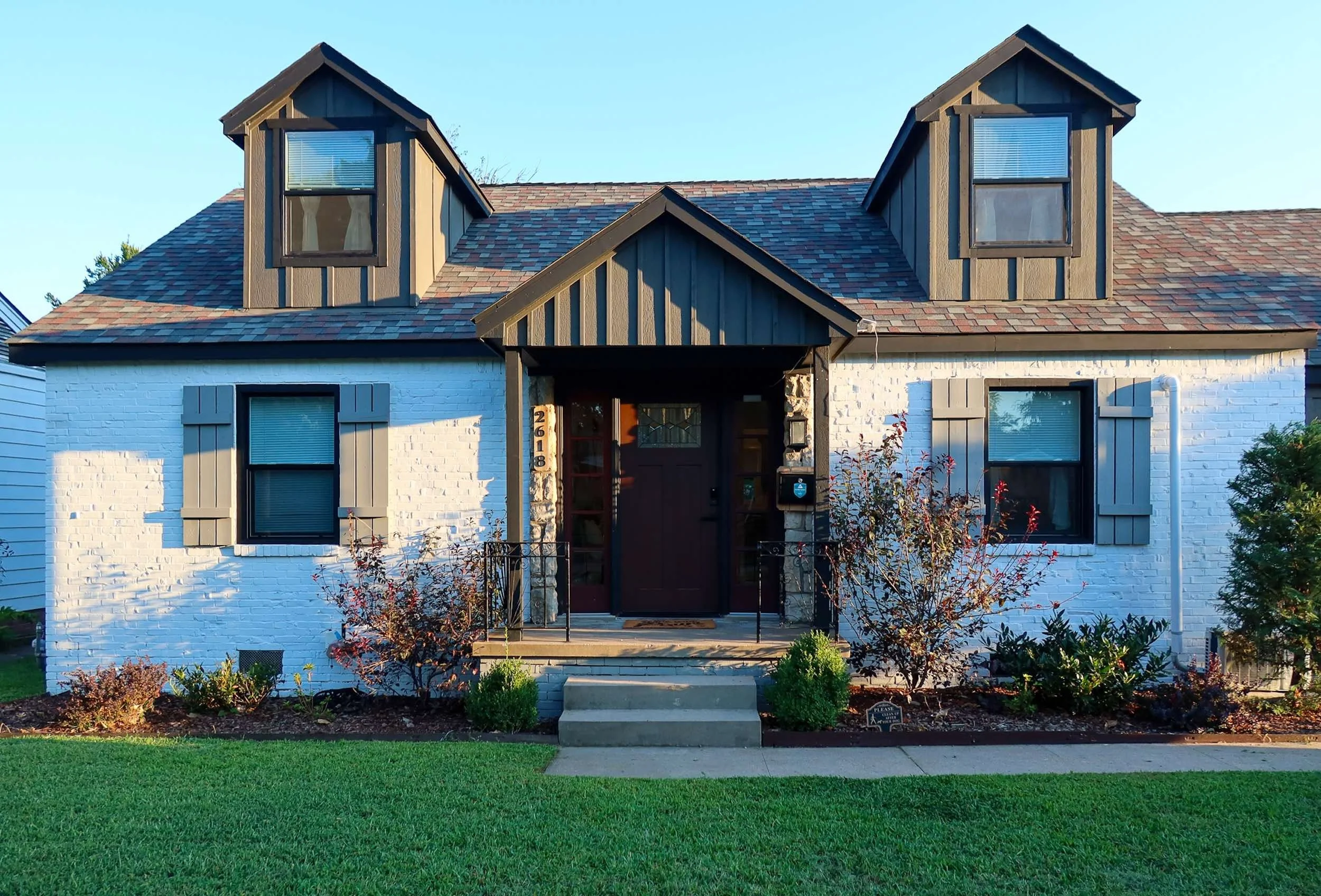 Front view of a white brick house with a dark brown front door and two dormer windows on the roof. The house has a small front porch with steps, surrounded by a green lawn and a garden with bushes and plants. The sky is clear.