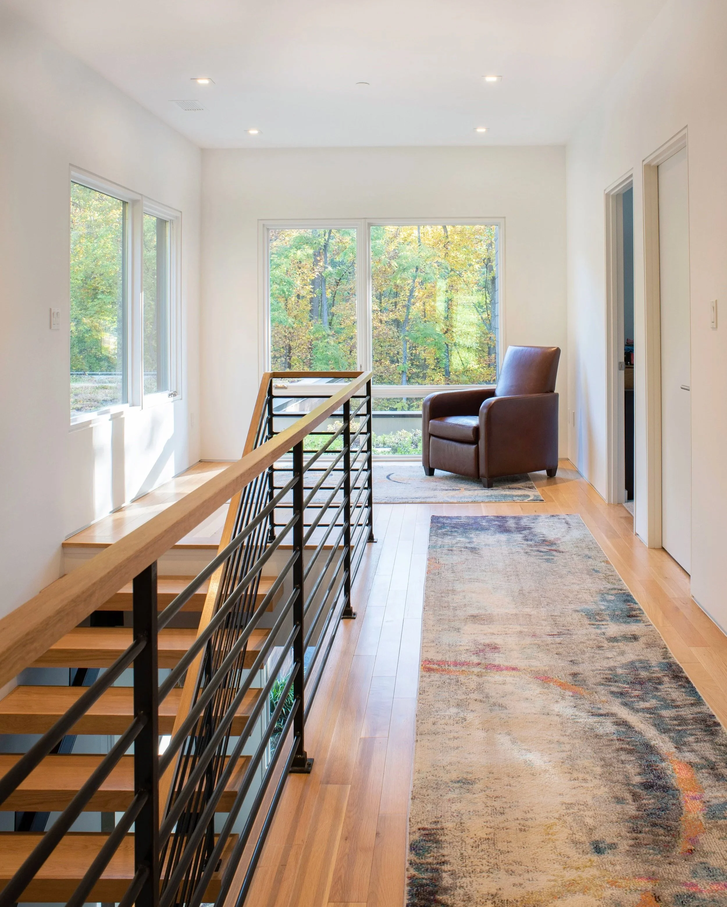 Inside view of a modern home hallway with large windows showing trees outside, a staircase with a wooden handrail, a brown armchair, a patterned rug, and white walls.