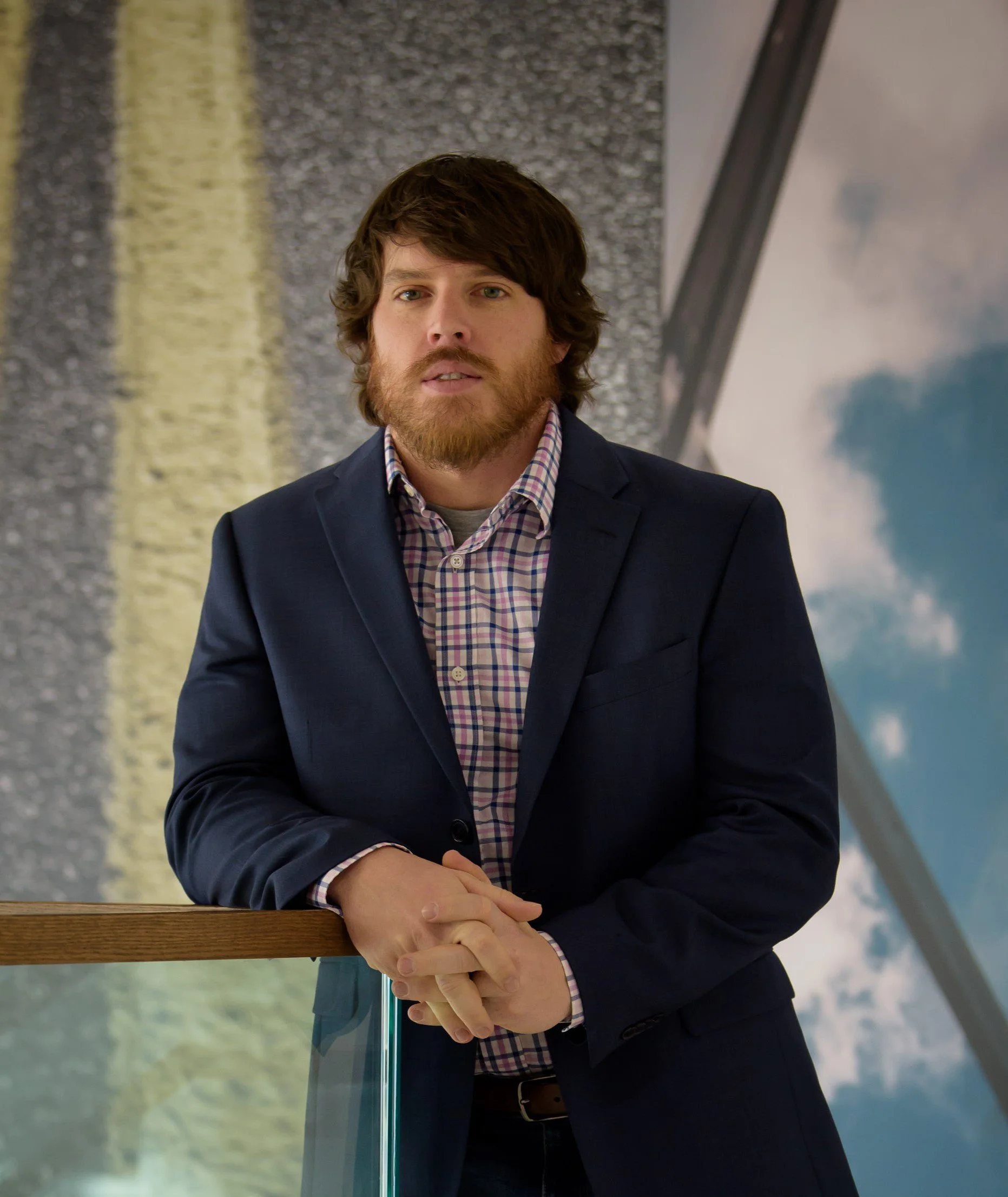 A man with brown hair and beard wearing a navy blazer and checked shirt, sitting with arms crossed, in front of a background with water and sky reflections.