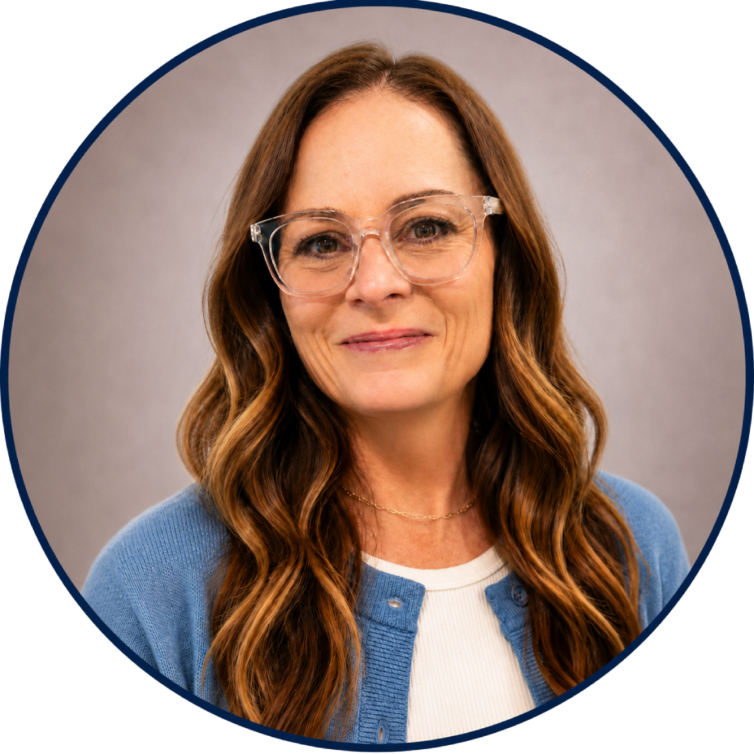 A woman with long wavy brown hair, wearing clear glasses, a white shirt, and a blue cardigan, smiling against a light gray backdrop.
