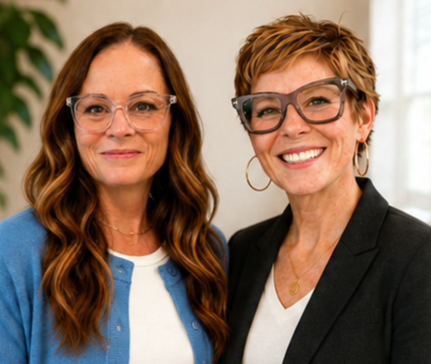 Two women smiling and posing together indoors, one with long wavy red hair and glasses, the other with short light brown hair and large dark glasses, both dressed casually.