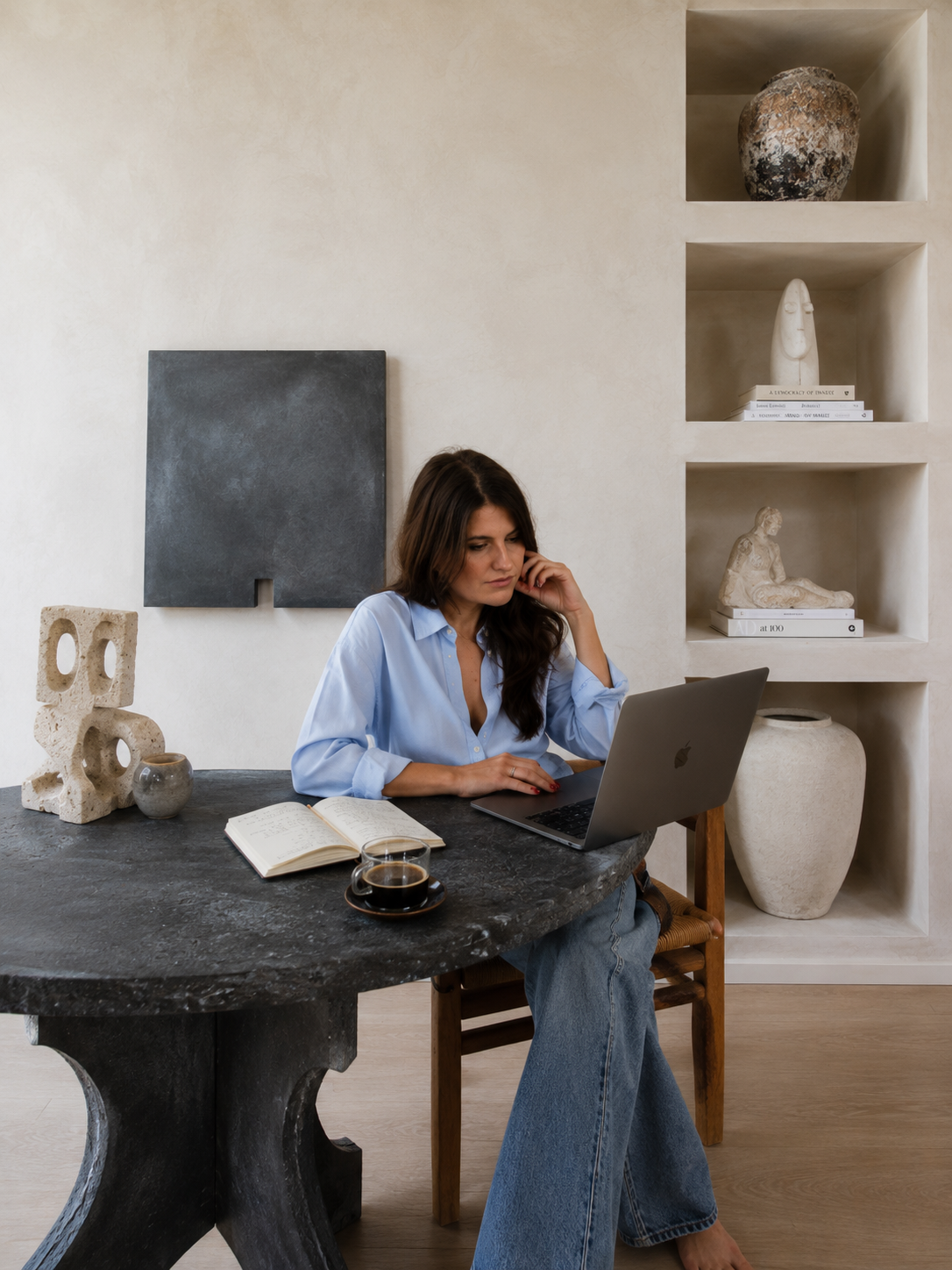 Une femme assise à une table en pierre, travaillant sur un ordinateur portable, avec un livre ouvert, une tasse de café et une petite tasse à côté. En arrière-plan, un mur beige avec des niches contenant des sculptures et des livres.