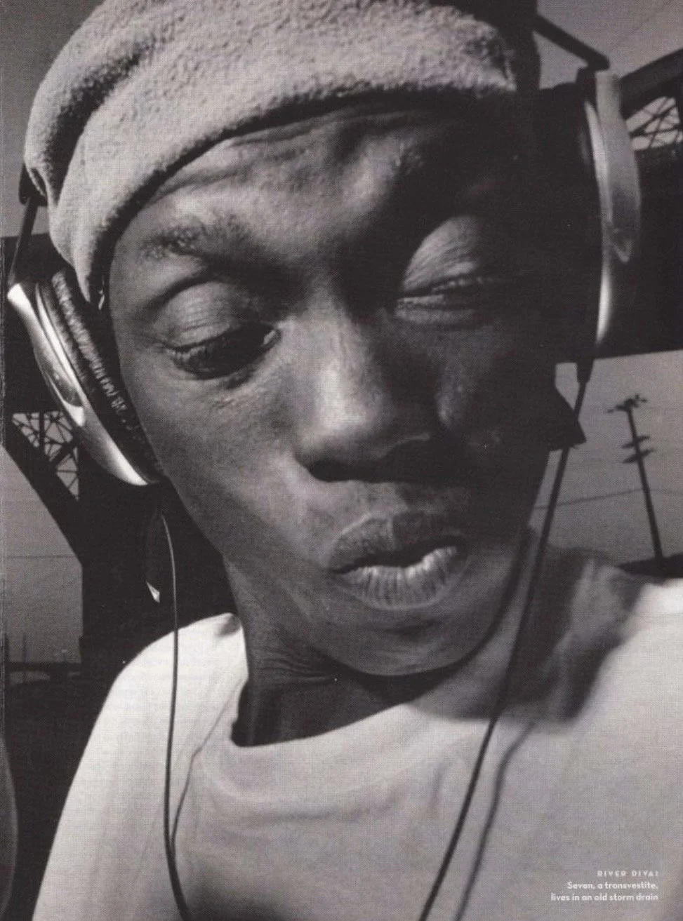 Close-up black-and-white photo of person wearing headphones and a beanie, with eyes closed and lips parted, outdoors with power lines and a tower in the background.