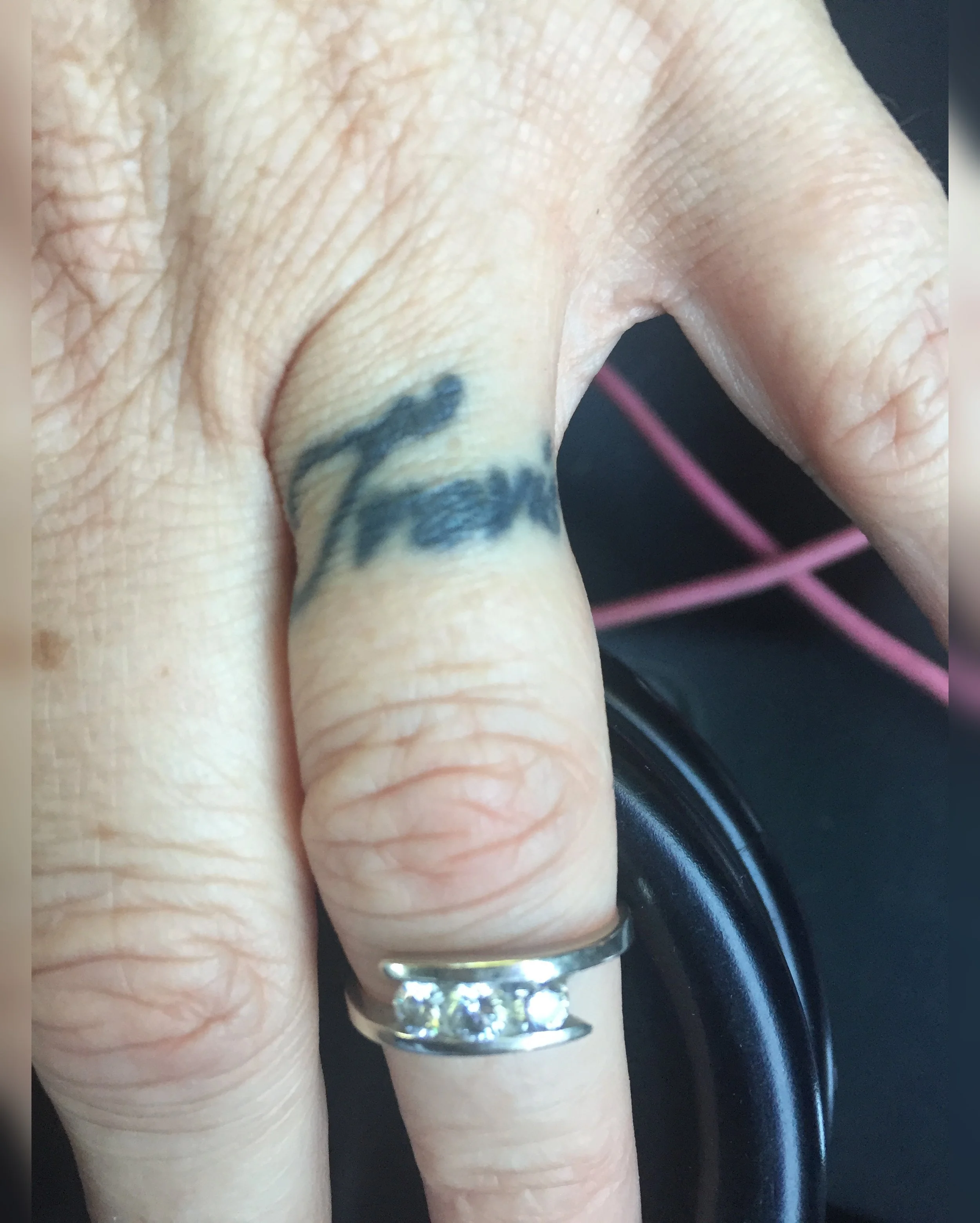 A close-up of a person's hand with a silver ring on the finger, holding a black steering wheel. The hand has visible wrinkles and a small tattoo or marking near the ring. Colorful reflections are visible on the ring.