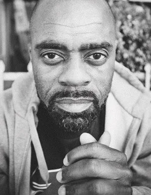 Close-up black and white photo of a man with a serious expression, looking into the camera, with a hand raised near his face.