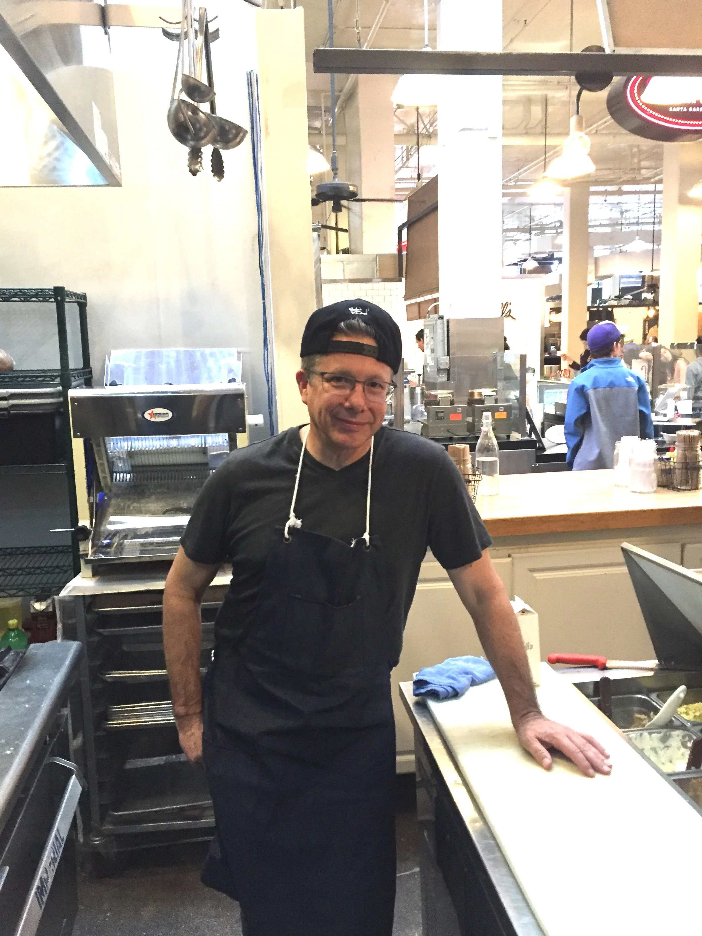 A man wearing glasses, a black cap backwards, a black shirt, and an apron standing in a busy restaurant kitchen, preparing food on a cutting board.