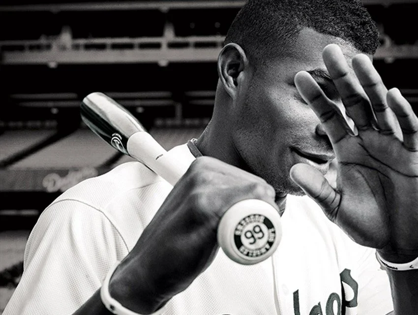 Close-up of a baseball player holding a bat over his shoulder, wearing a uniform, with one hand raised partially covering his face and a serious expression
