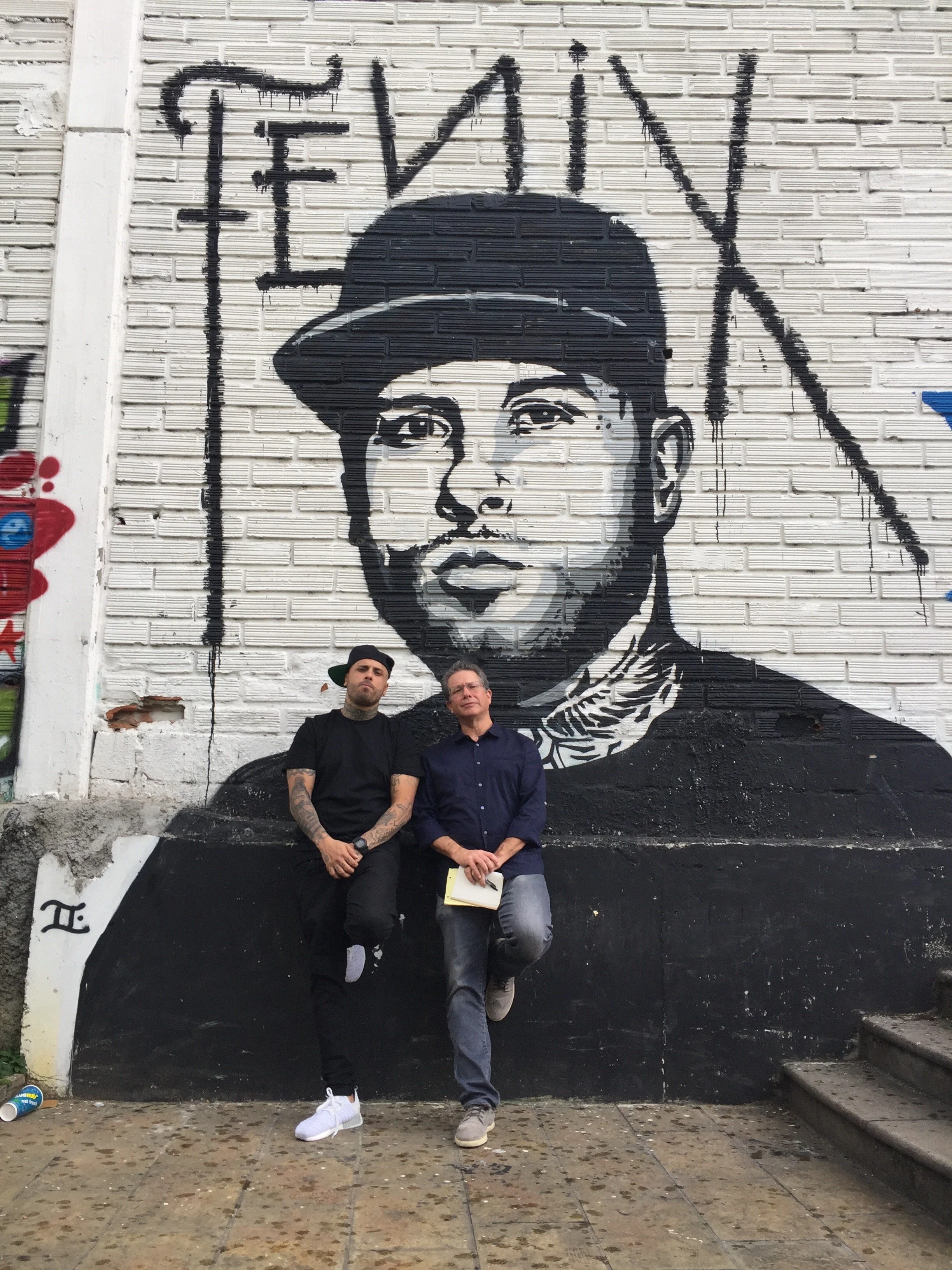 Two men standing in front of a large black and white mural of a person's face on a brick wall, with the word 'FEAR' at the top.