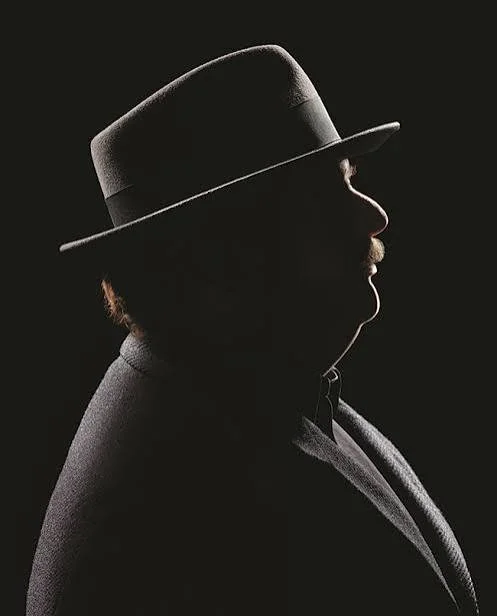 Silhouette of Pulitzer-winning food critic Jonathan Gold wearing a fedora hat and a suit against a dark background.