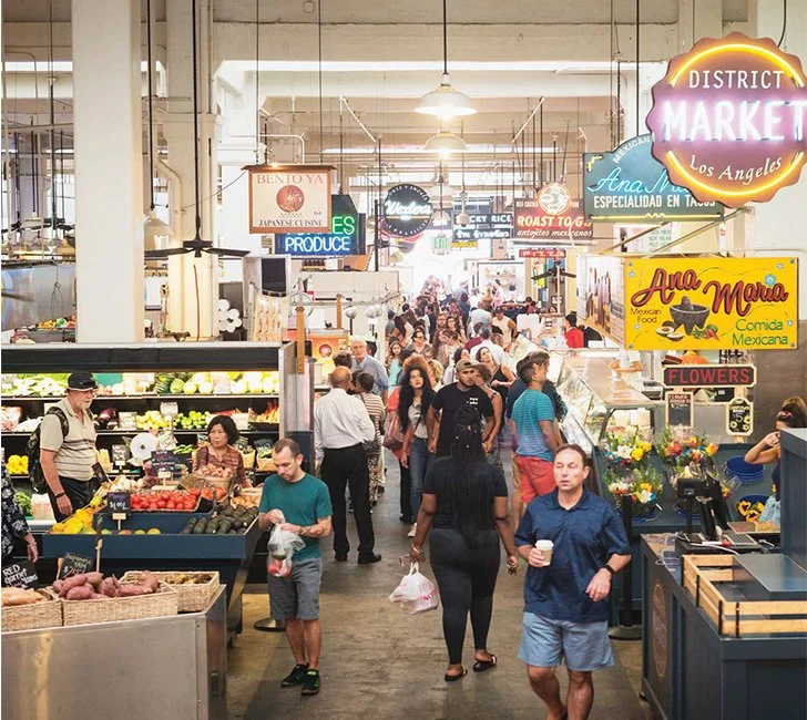 Inside a busy indoor marketplace with various food stalls and signs, many shoppers walking around and purchasing items.