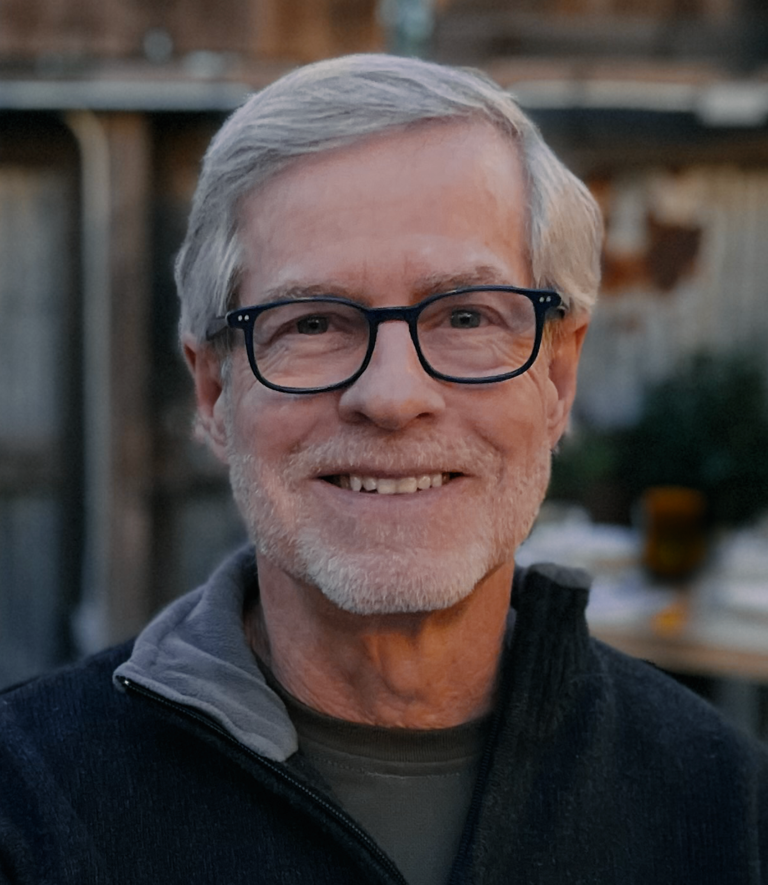 Close-up of an elderly man with gray hair, wearing glasses, smiling, dressed in a gray hoodie and a black fleece jacket, outdoors in a natural setting.