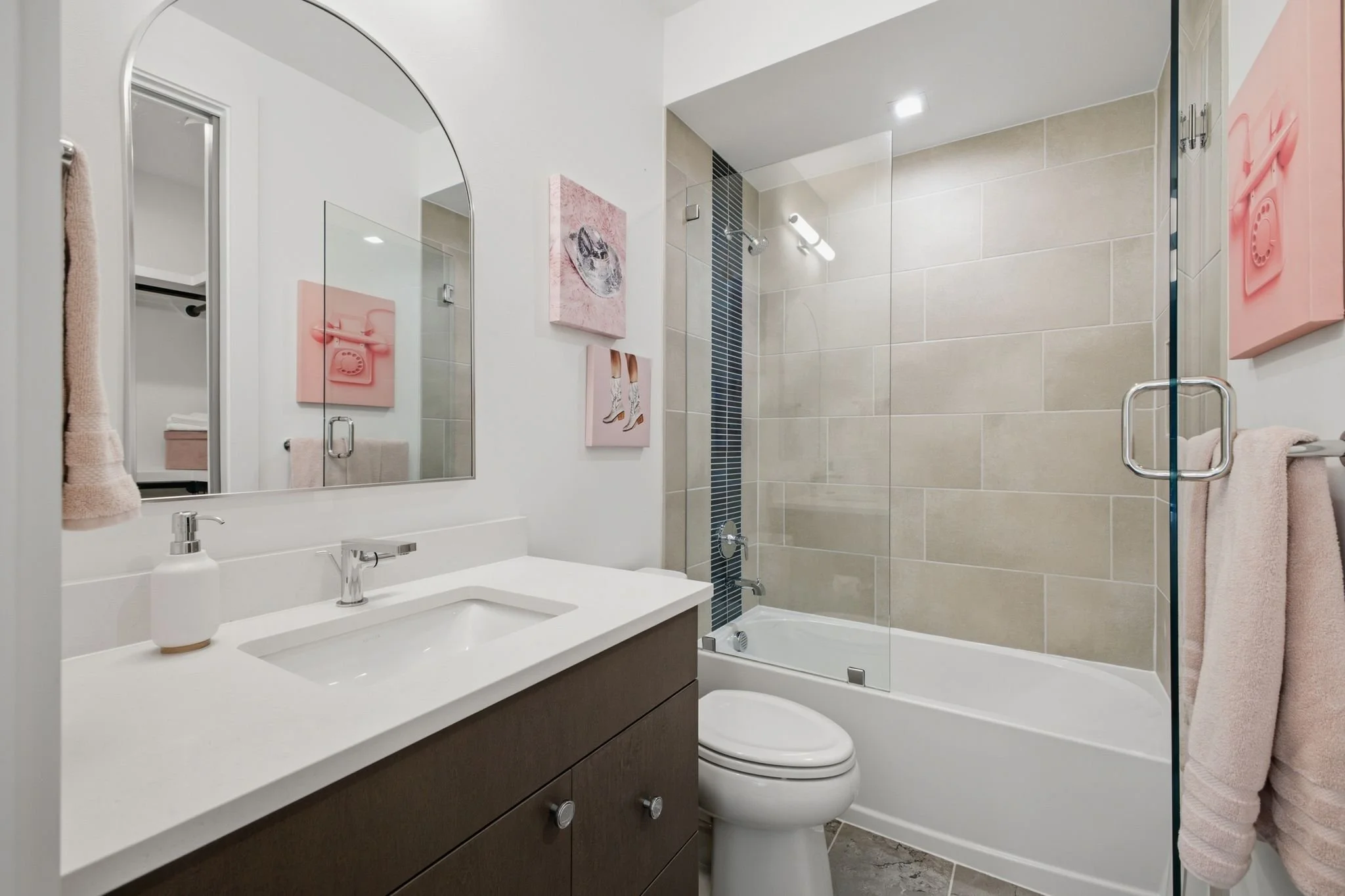 Modern bathroom with vanity sink, large mirror, artwork on the wall, toilet, bathtub with glass shower door, beige tiled walls, and pink towels.