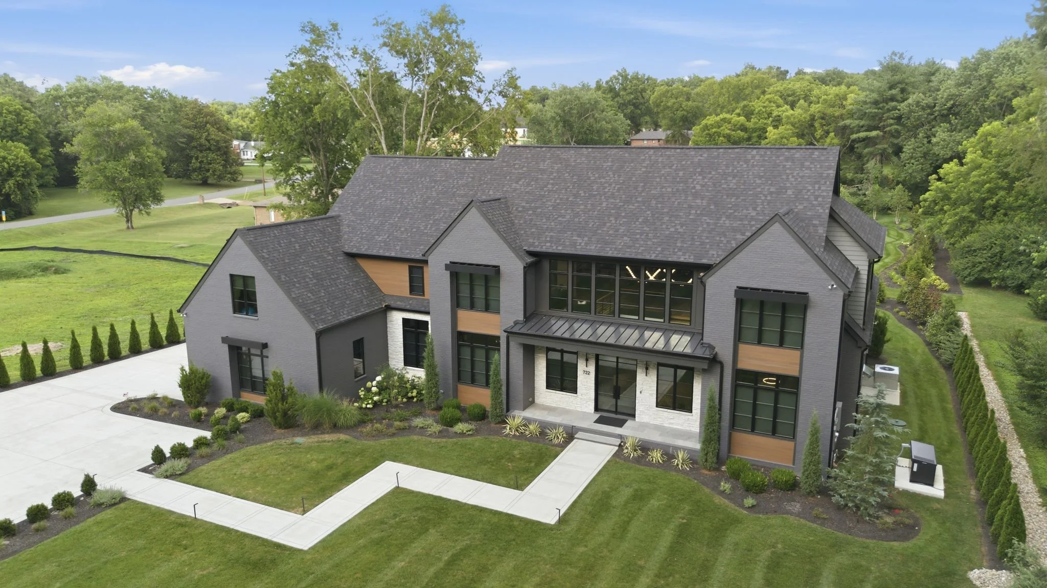 A modern two-story house with grey brick exterior, black window frames, and a landscaped front yard with small shrubs and trees, surrounded by green lawns and trees under a clear blue sky.
