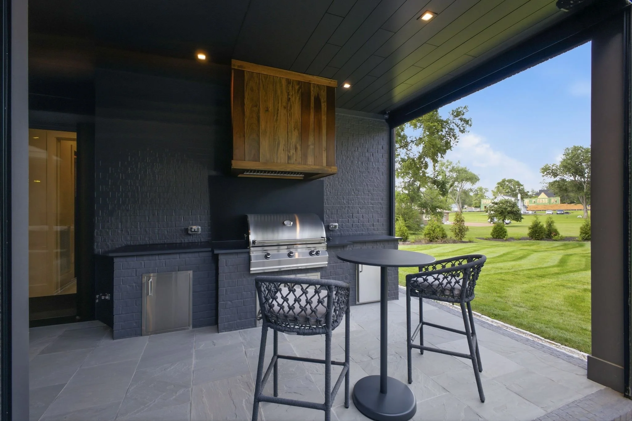 Outdoor covered patio area with a black brick wall, stainless steel grill, wooden cabinet, and a small round table with chairs. Green lawn and trees are visible in the background under a clear blue sky.