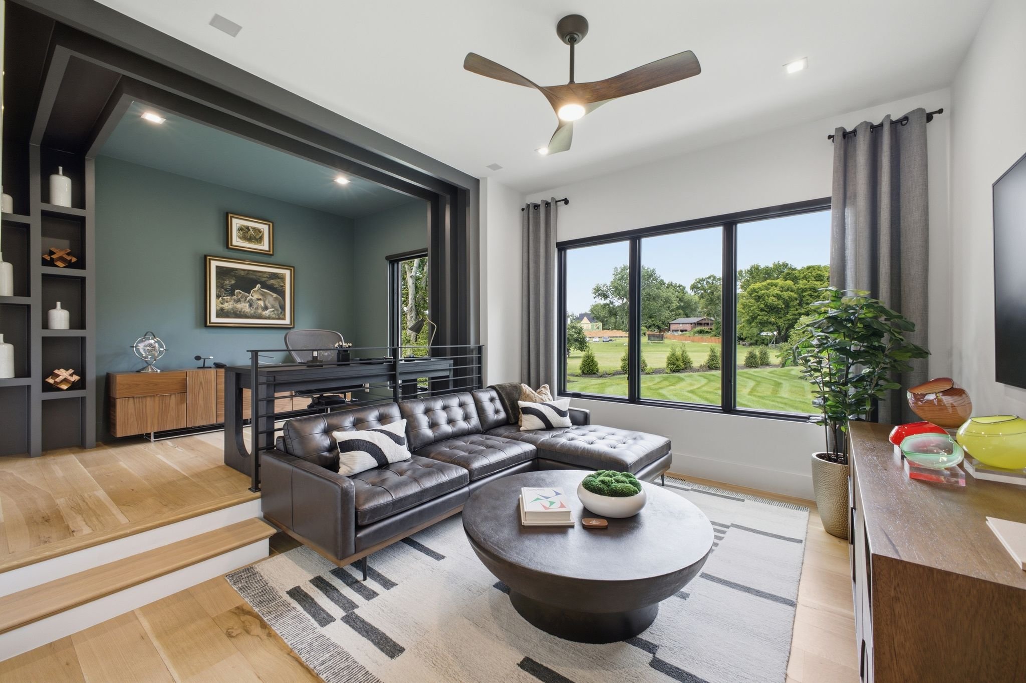 A modern living room with large windows showing a suburban yard. The room features a black leather sectional sofa, a wooden coffee table with decorative bowls, a small round carpet, and various decorative items. There's a ceiling fan with wooden blad