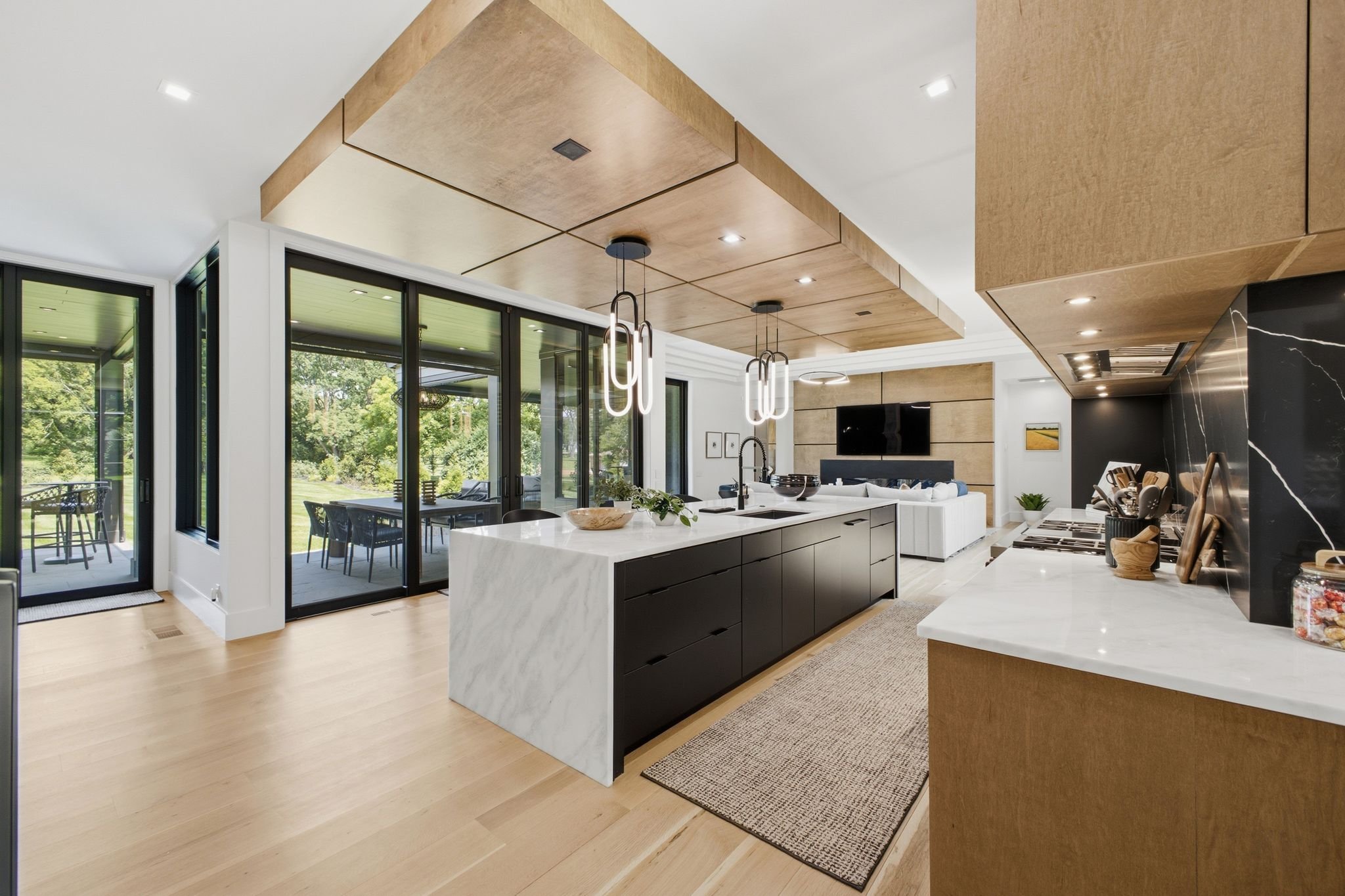 Modern kitchen with white island, black cabinets, wooden ceiling panels, pendant lights, large sliding glass doors leading to patio, view of greenery outside, and light wood flooring.