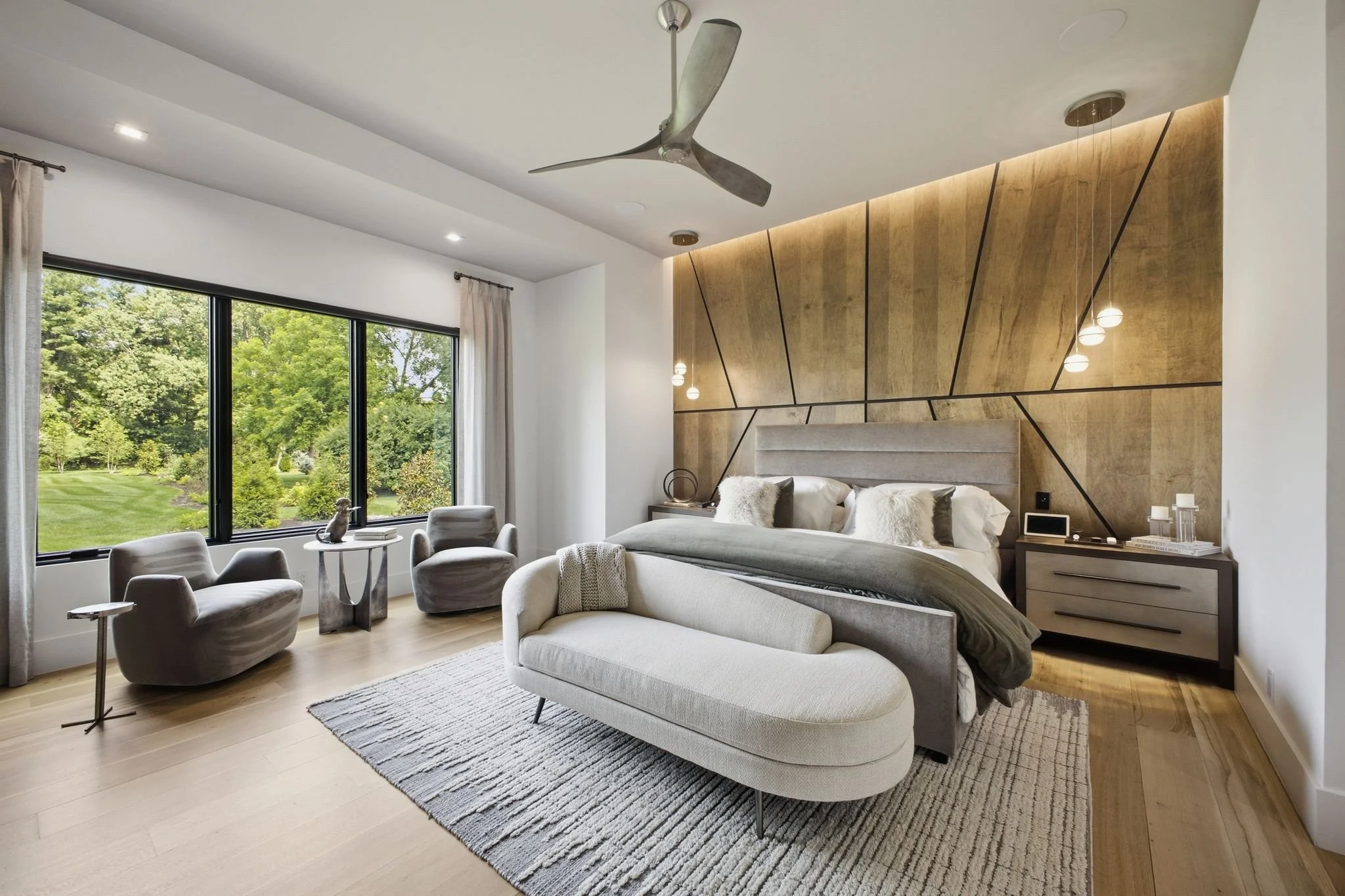 Modern bedroom with white and beige furniture, large window showing greenery outside, and decorative lighting fixtures.