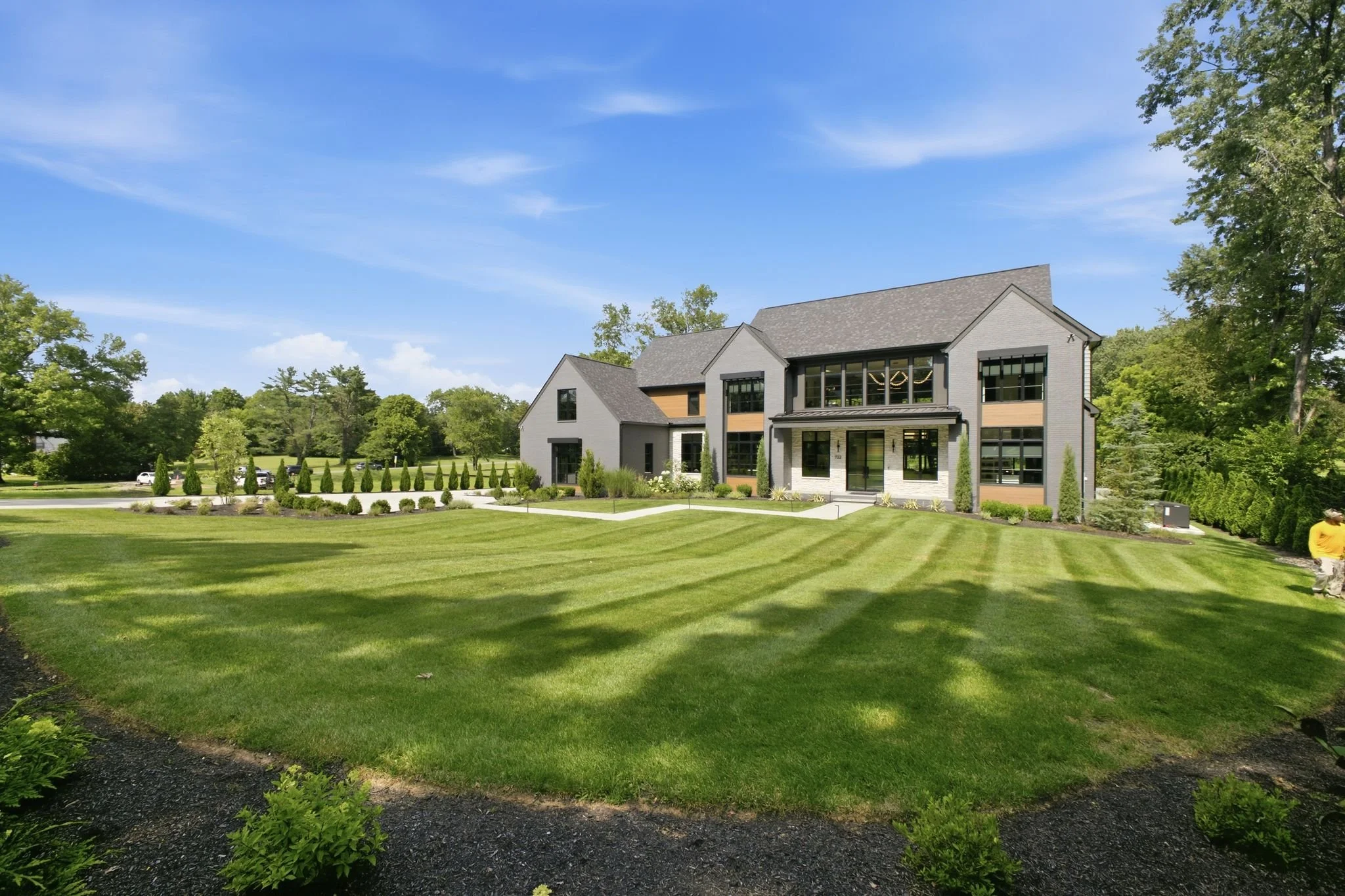 Modern two-story house with large windows and a gray roof, surrounded by green landscaping and a well-maintained lawn, with tall trees and a blue sky in the background.