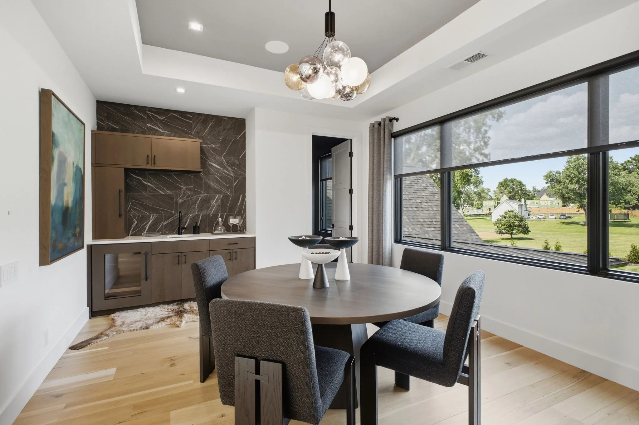 Contemporary dining room with a round wooden table, six upholstered chairs, a modern chandelier with globes, large windows with gray curtains, and a view of a suburban yard with trees and houses.