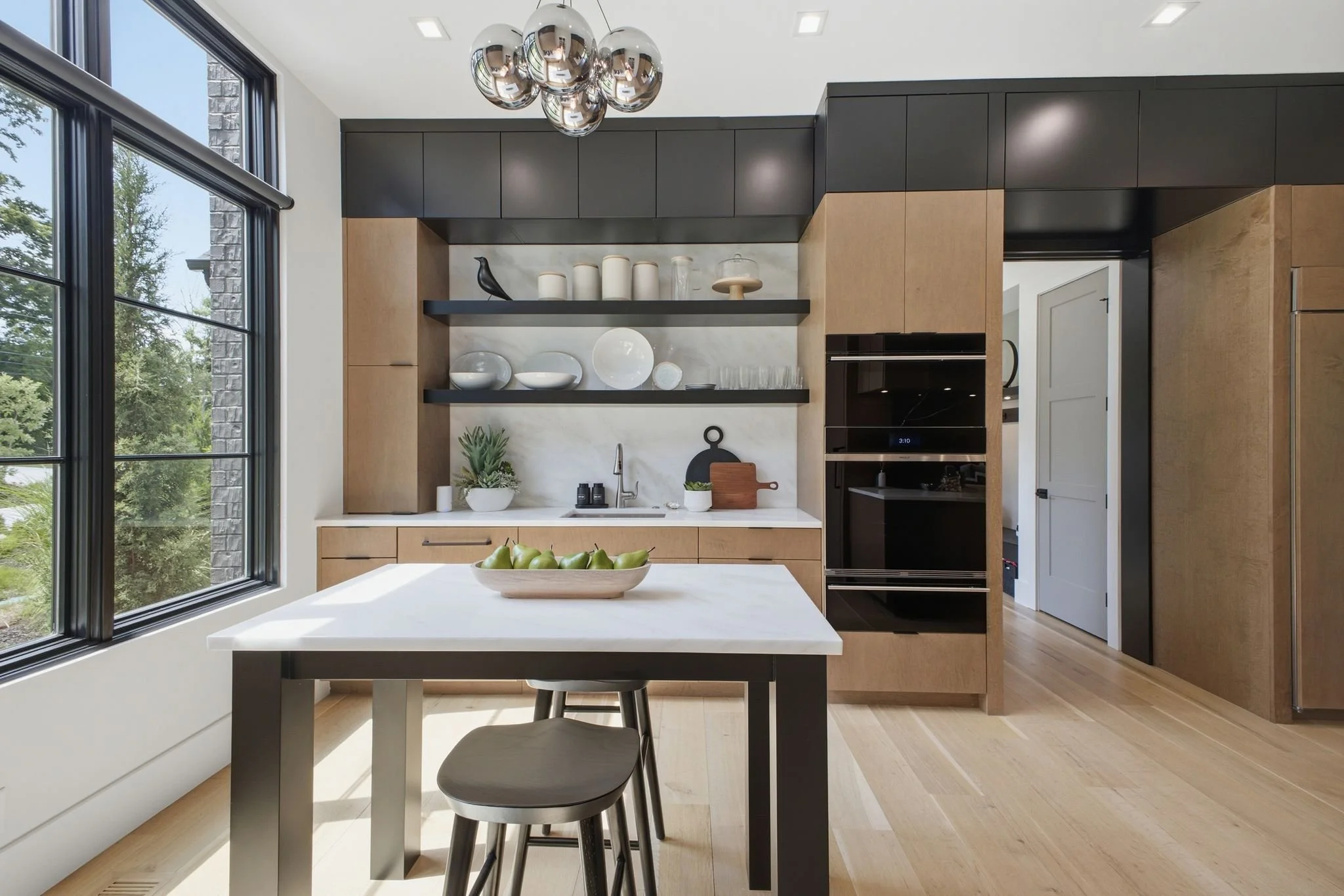 A modern kitchen with large windows, a white island with green apples in a bowl, black and natural wood cabinetry, open shelving with white and glass decor, a black built-in oven, and a contemporary chandelier with reflective chrome balls.