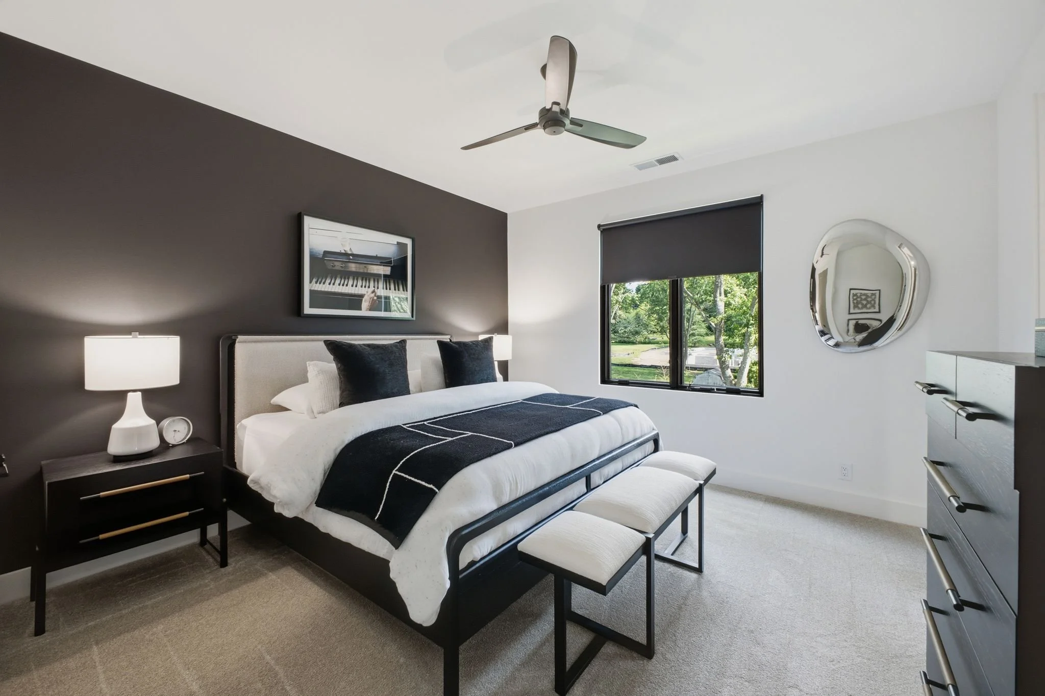 Modern bedroom with a bed, nightstand, lamp, and window showing greenery outside.