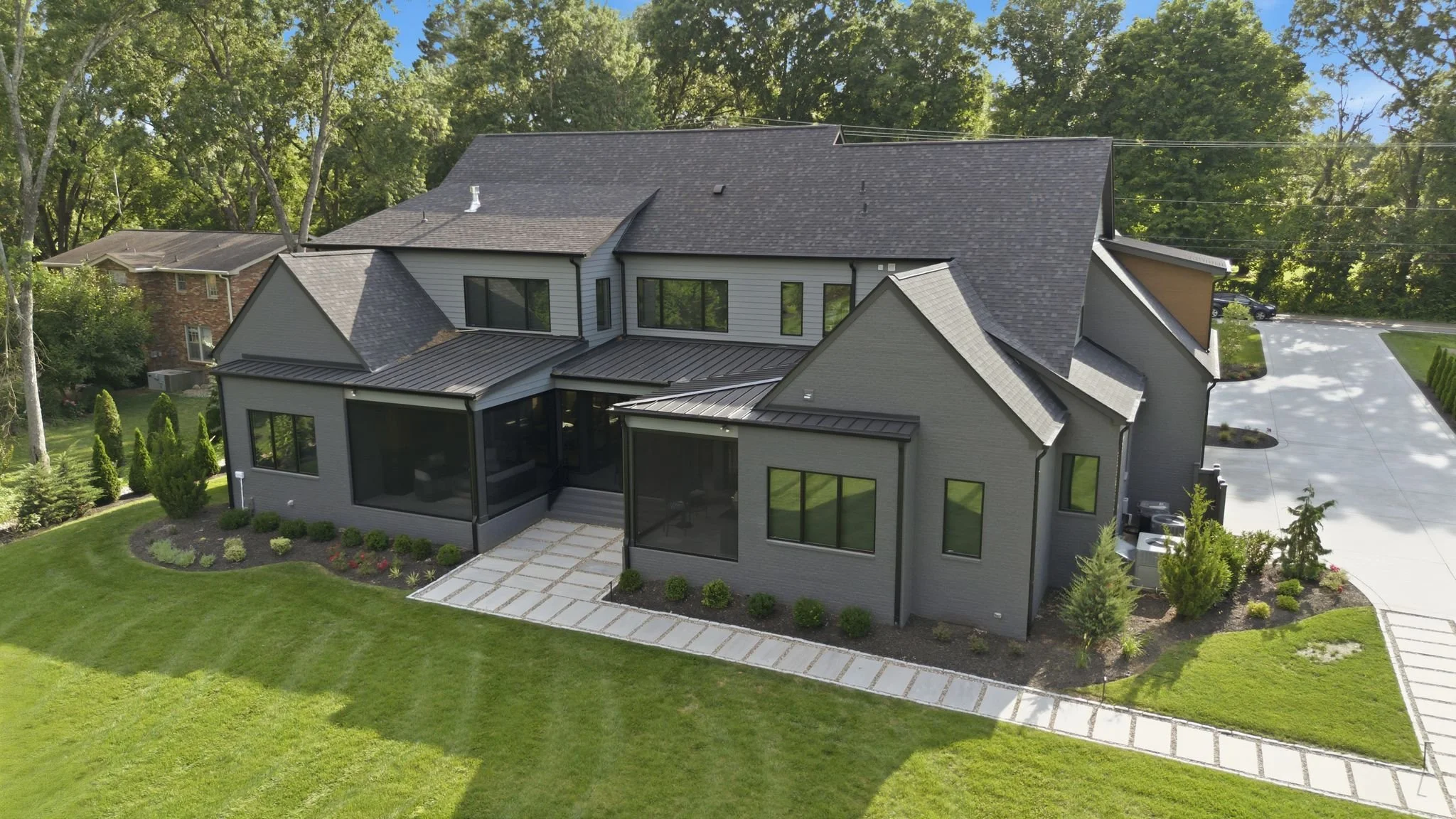 A modern two-story house with gray exterior walls, multiple gable roofs, large windows, a screened porch, and a landscaped front yard with a lawn and garden beds, surrounded by green trees.