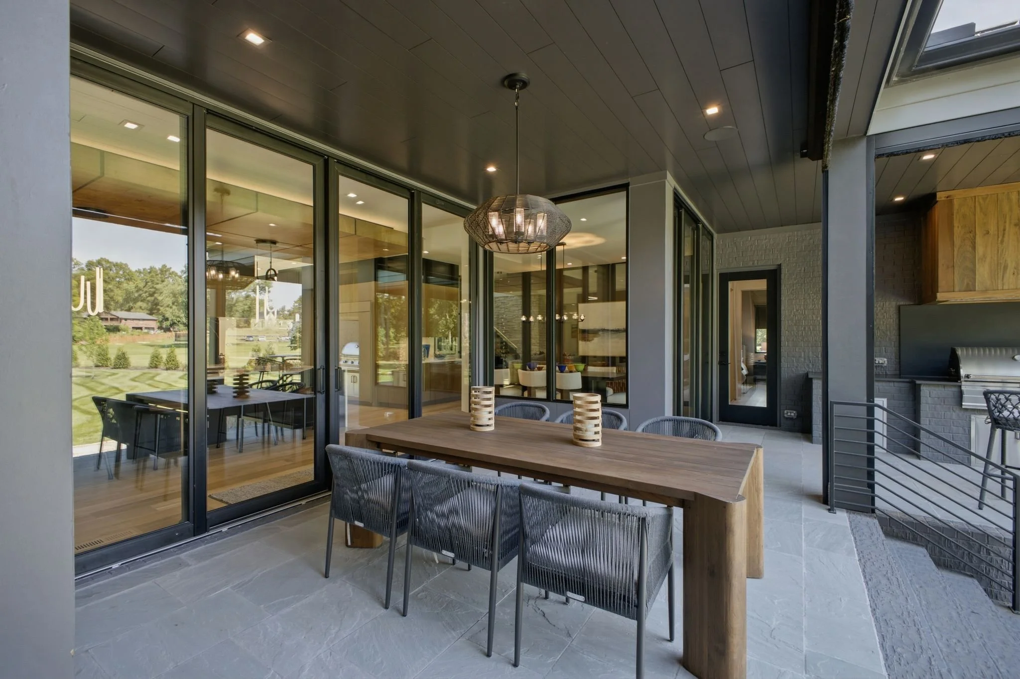 Outdoor patio with wooden dining table, metal chairs, hanging pendant light, glass sliding doors, and view of a golf course in the background.