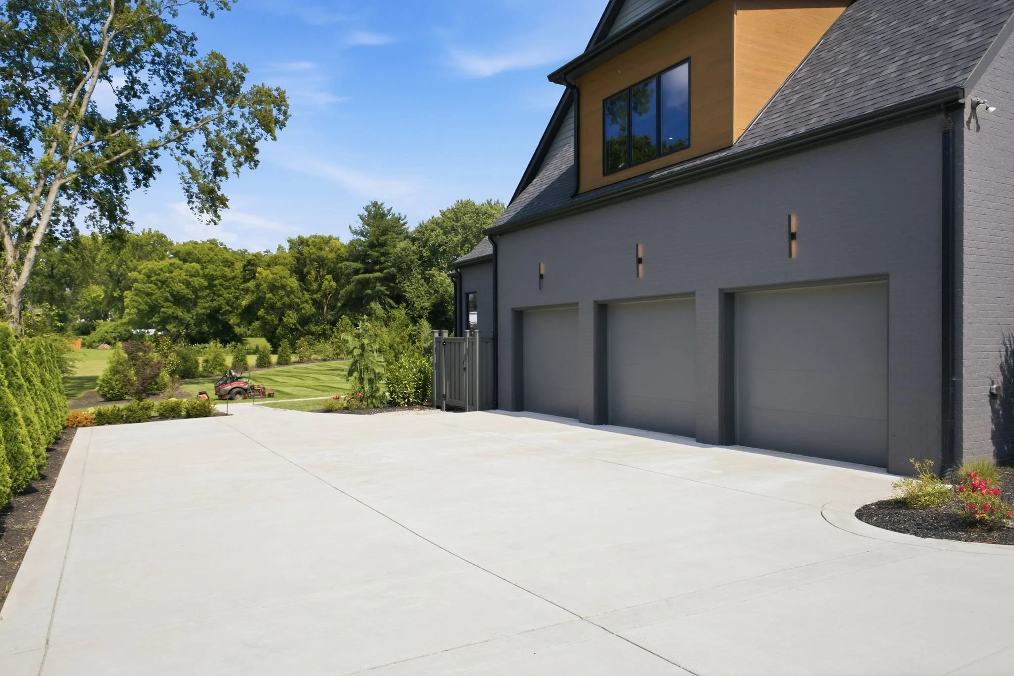 Modern house with gray brick exterior, large windows, and black roofing. Clean concrete driveway and landscaped yard with trees and shrubs under a clear blue sky.