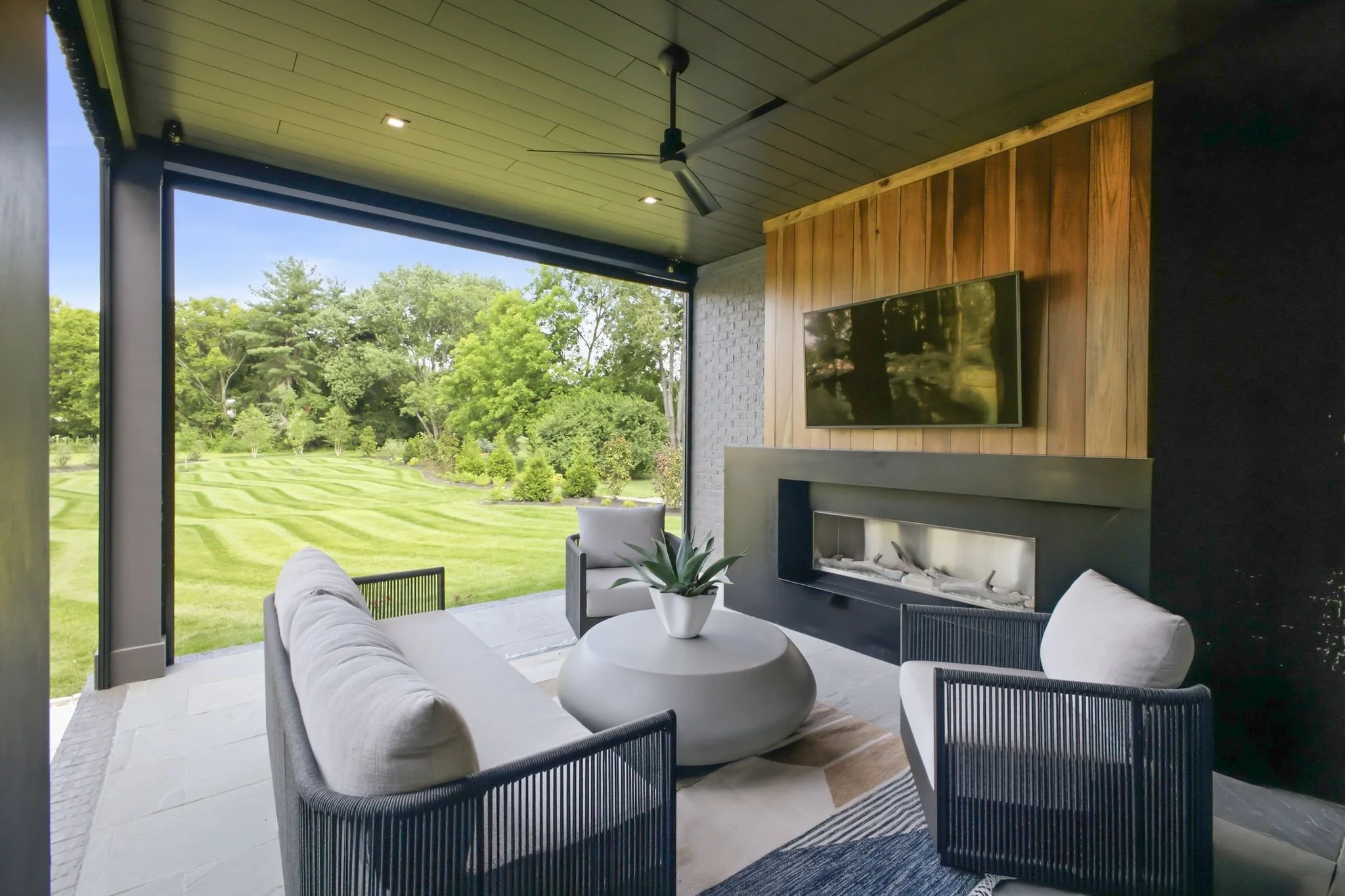 Covered outdoor patio with seating area, fireplace, wall-mounted TV, ceiling fan, and views of a lush green backyard with trees.