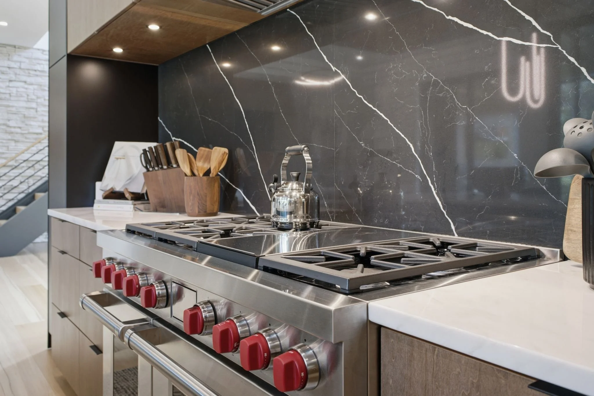 Modern kitchen with black marble backsplash, stainless steel stove with red control knobs, a kettle on top, and wooden utensils in a container on the countertop.