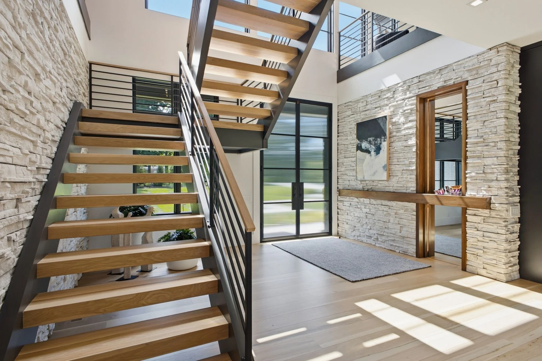 Modern interior staircase with wooden steps, metal support, and black railing inside a house with stone and painted walls, large windows, and a front door near the entrance.