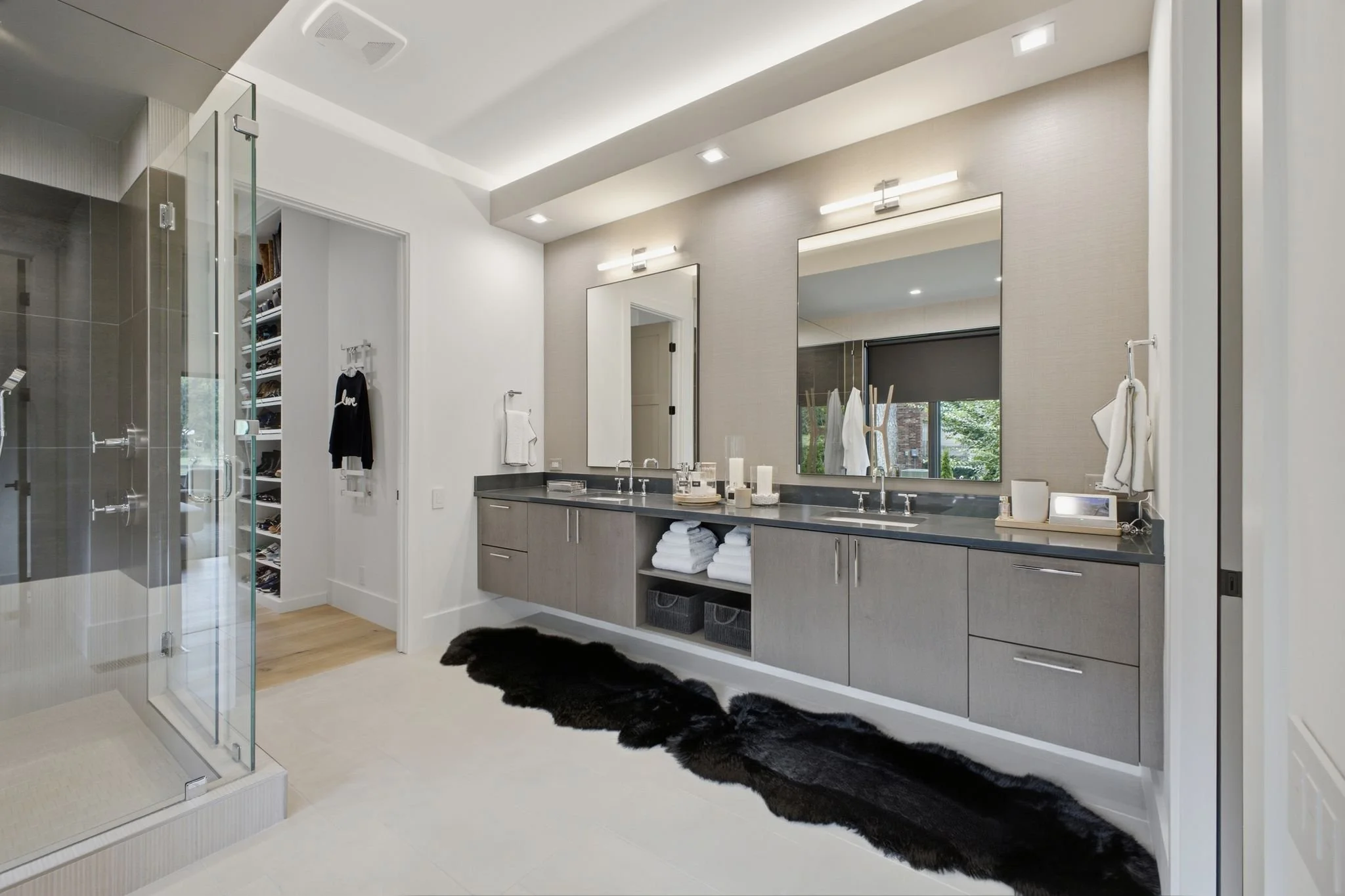 Modern bathroom with dual sinks, large mirrors, and a shower stall with glass door. There are towels neatly stored below the sinks, and decorative candles and toiletries on the countertop. A window with a view of trees is reflected in the mirror.