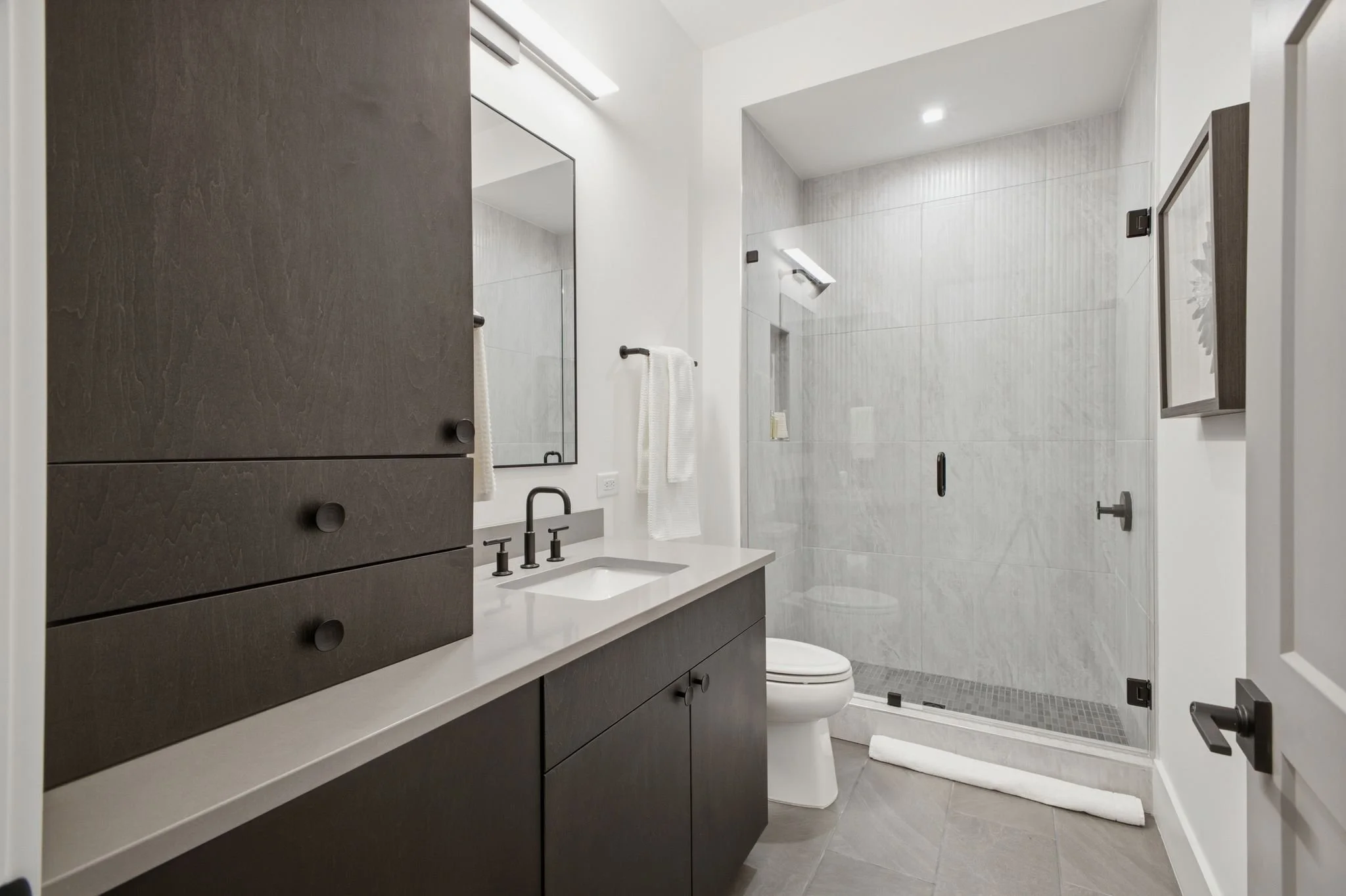 Modern bathroom with dark vanity, white countertop, black fixtures, large mirror, white towels, and a glass-enclosed shower with gray tile walls and a rain shower head.