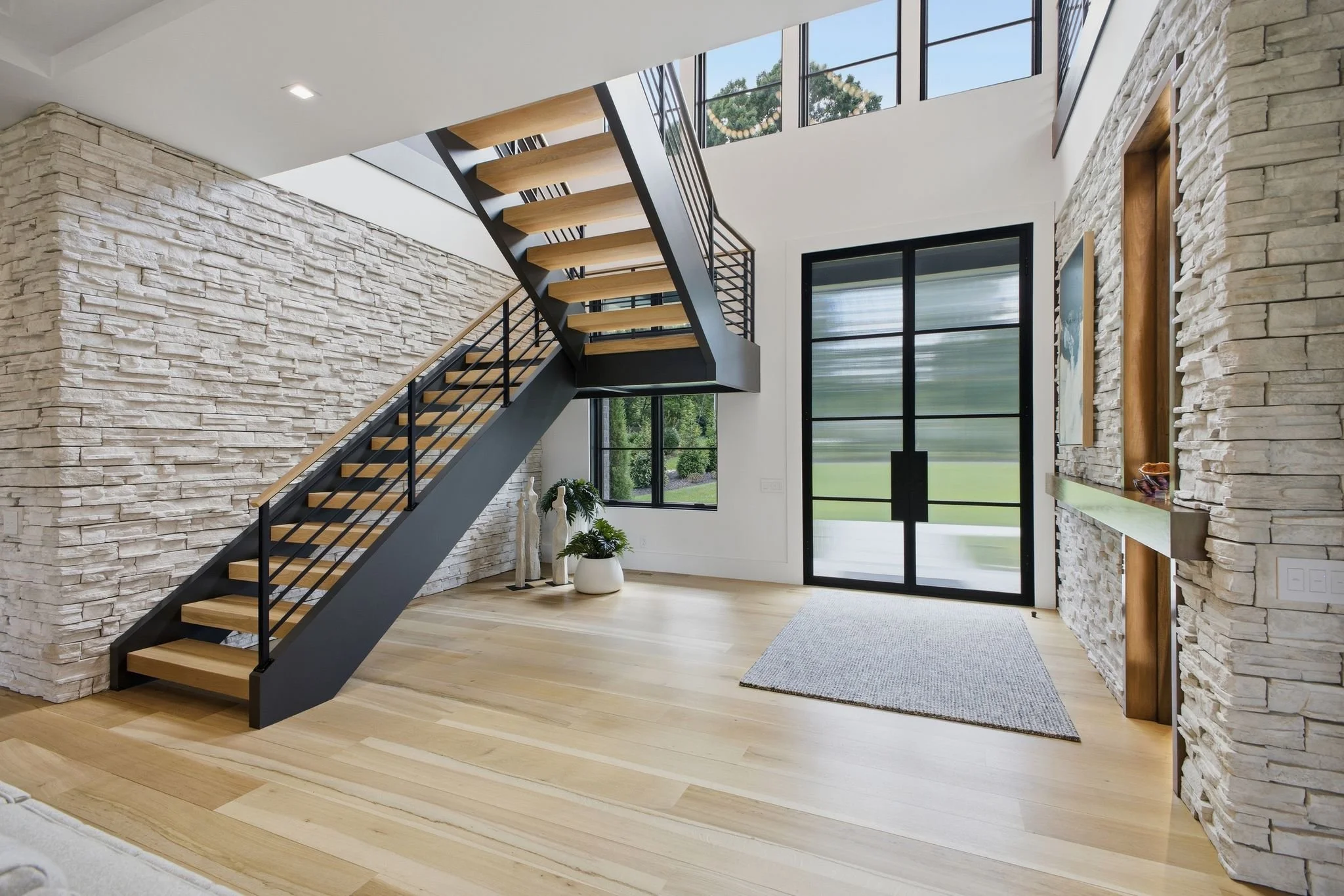 Interior of a modern house with a staircase, stone accent walls, large windows, and hardwood floors.