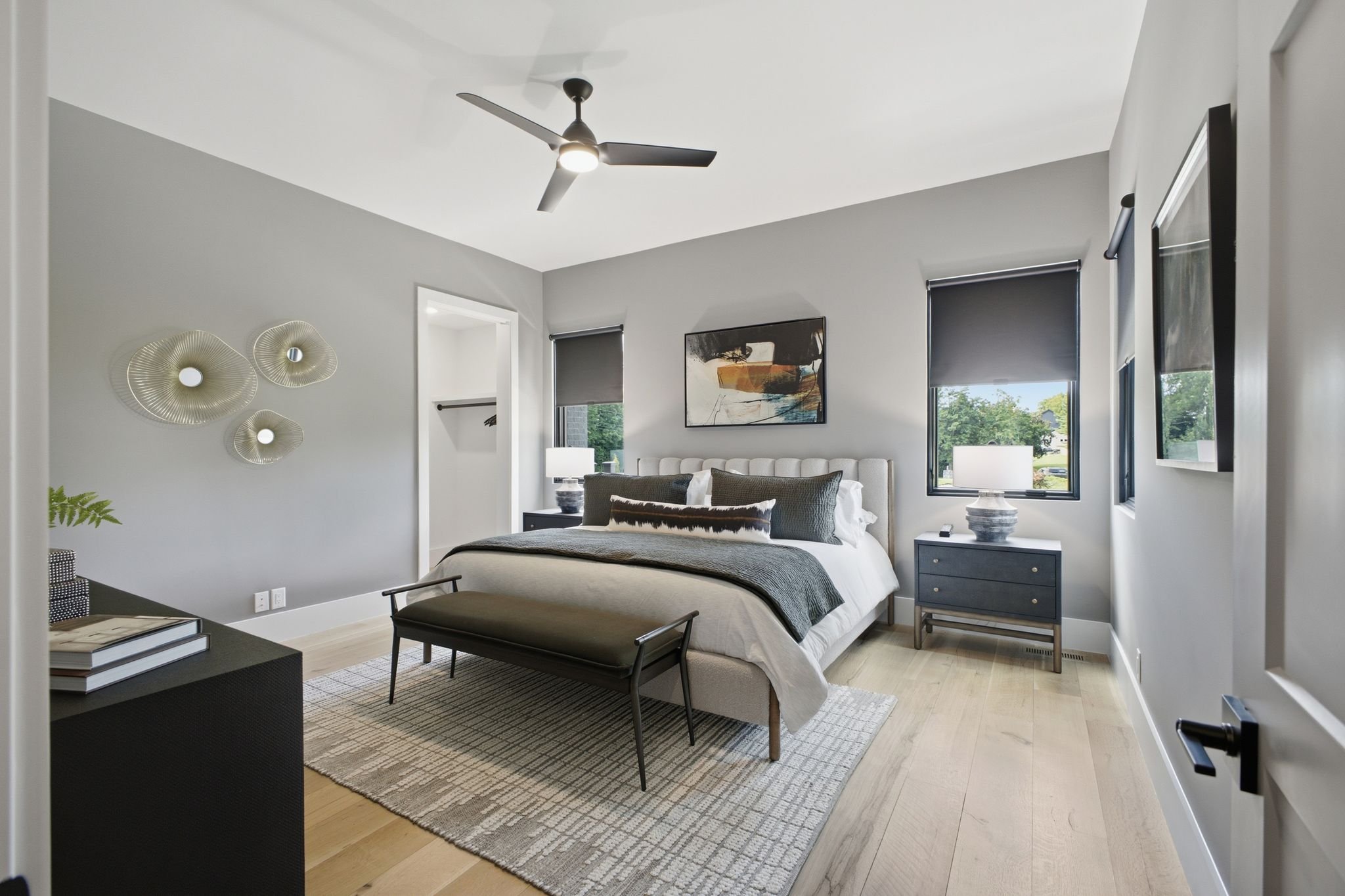 A modern bedroom with a bed, nightstands, lamps, and wall art, featuring gray walls and large windows with black blinds.