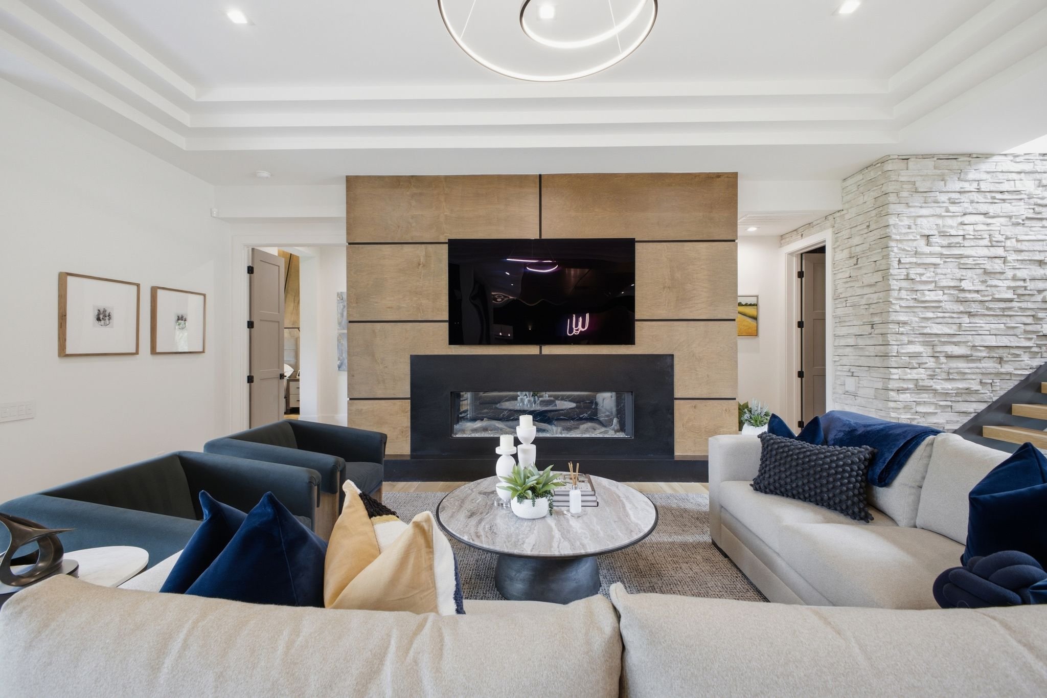 Modern living room with a white sofa, dark armchair, and a round coffee table with decorative items, a fireplace, a large flat-screen TV, and a textured stone accent wall.
