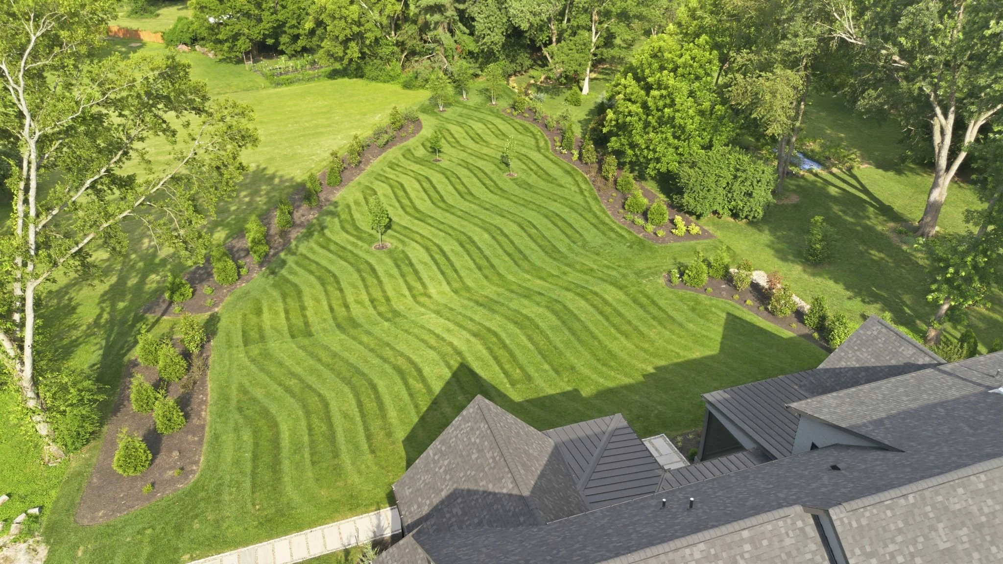 A backyard with neatly mowed grass in a striped pattern, lined with young trees and flowerbeds, surrounded by mature trees and a house with multiple rooftops.