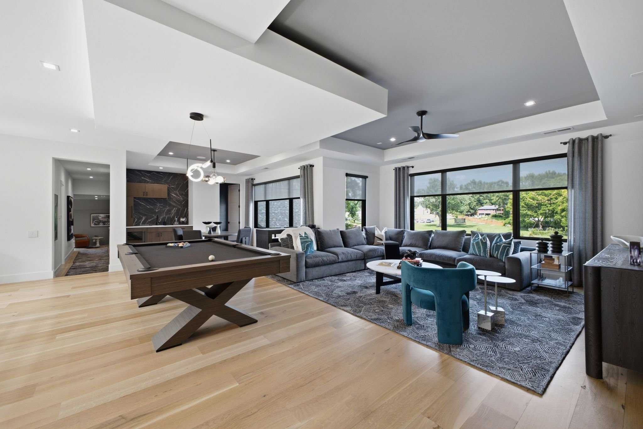 Modern living room with light wood flooring, a gray sectional sofa, large windows with gray curtains, and a ceiling fan. There is a pool table in the foreground, and a kitchen area visible in the background.