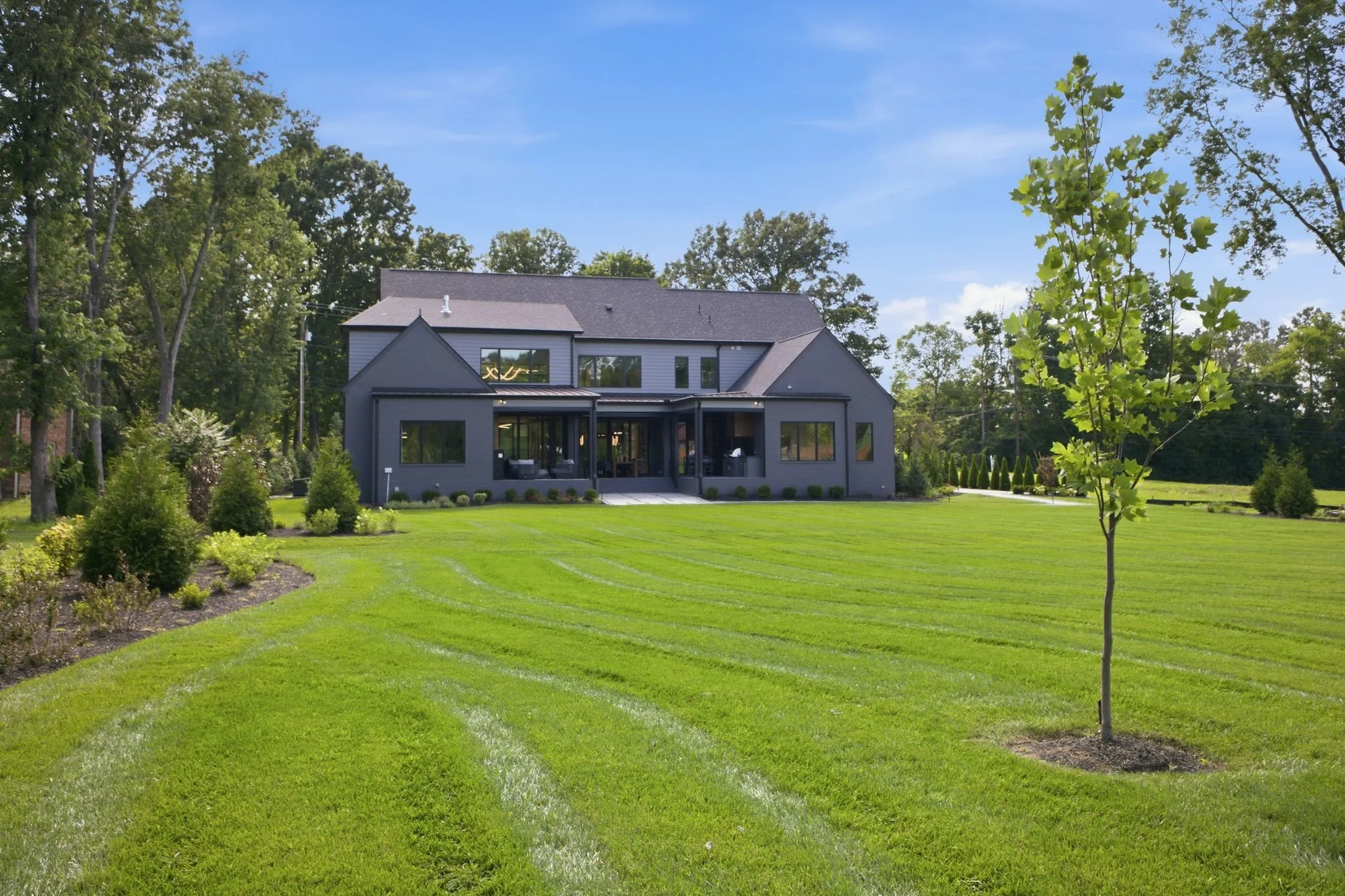 A modern gray house with large windows set in a lush green lawn with young trees and shrubs, surrounded by tall trees under a blue sky.
